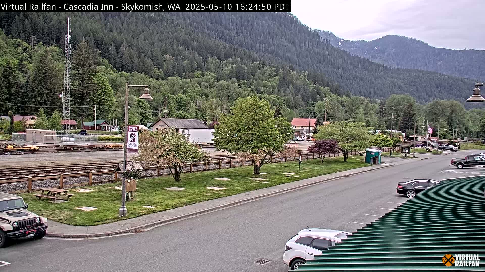 A partly cloudy day reveals a view of a small town nestled in a valley, with a train track, cars parked along a road, and lush green trees and mountains in the background.