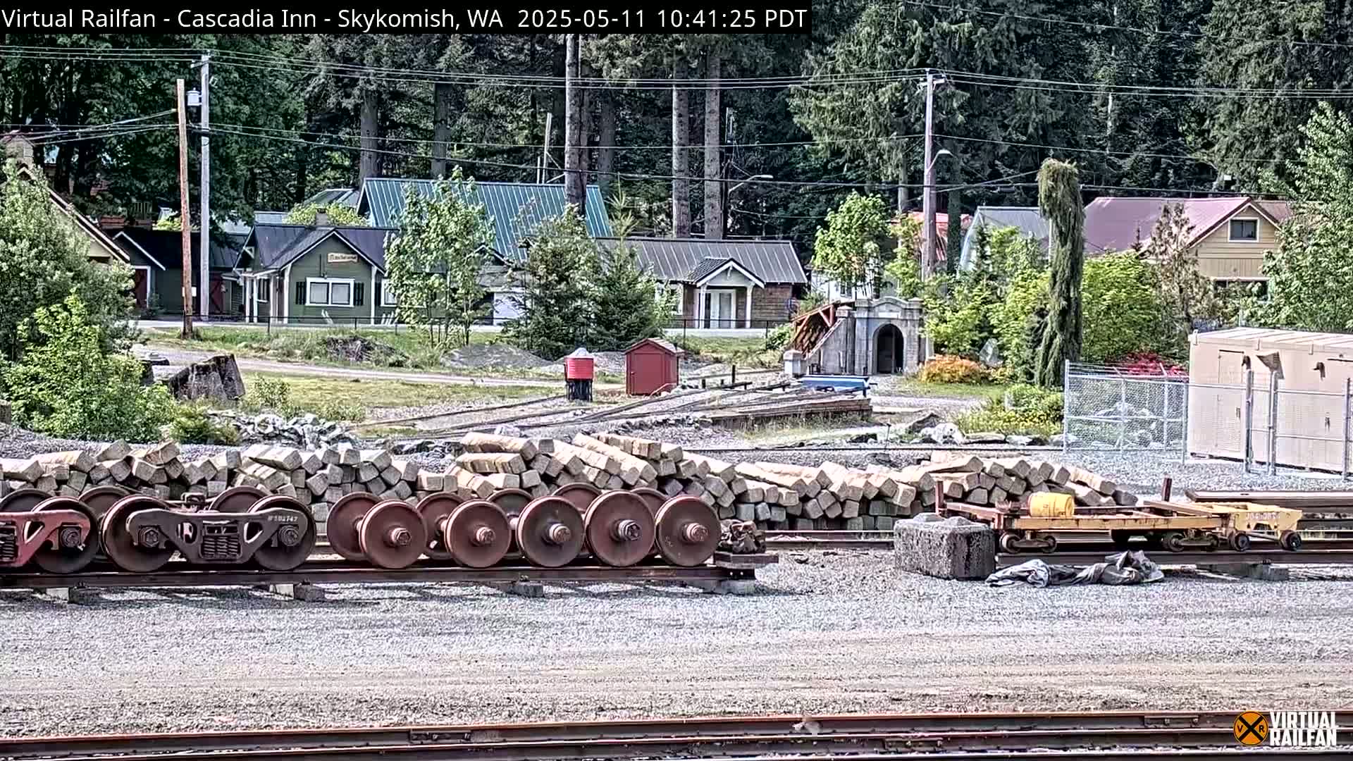 A sunny day shows train wheels and ties stacked near a railway,  with small houses and trees in the background.