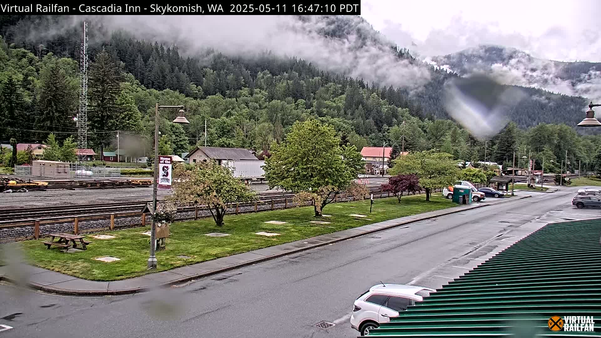A rain-slicked street and parking lot sit before a railroad track and grassy area, all nestled in a valley with a misty, forested mountainside rising in the background.