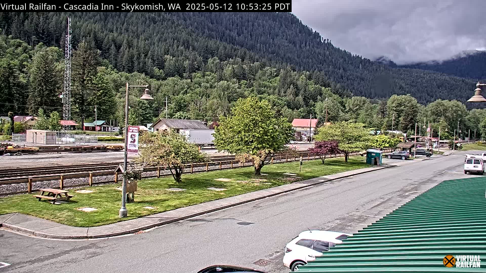 A partly cloudy day reveals a small town nestled beside a mountain range, featuring train tracks, a parking lot, and several cars.
