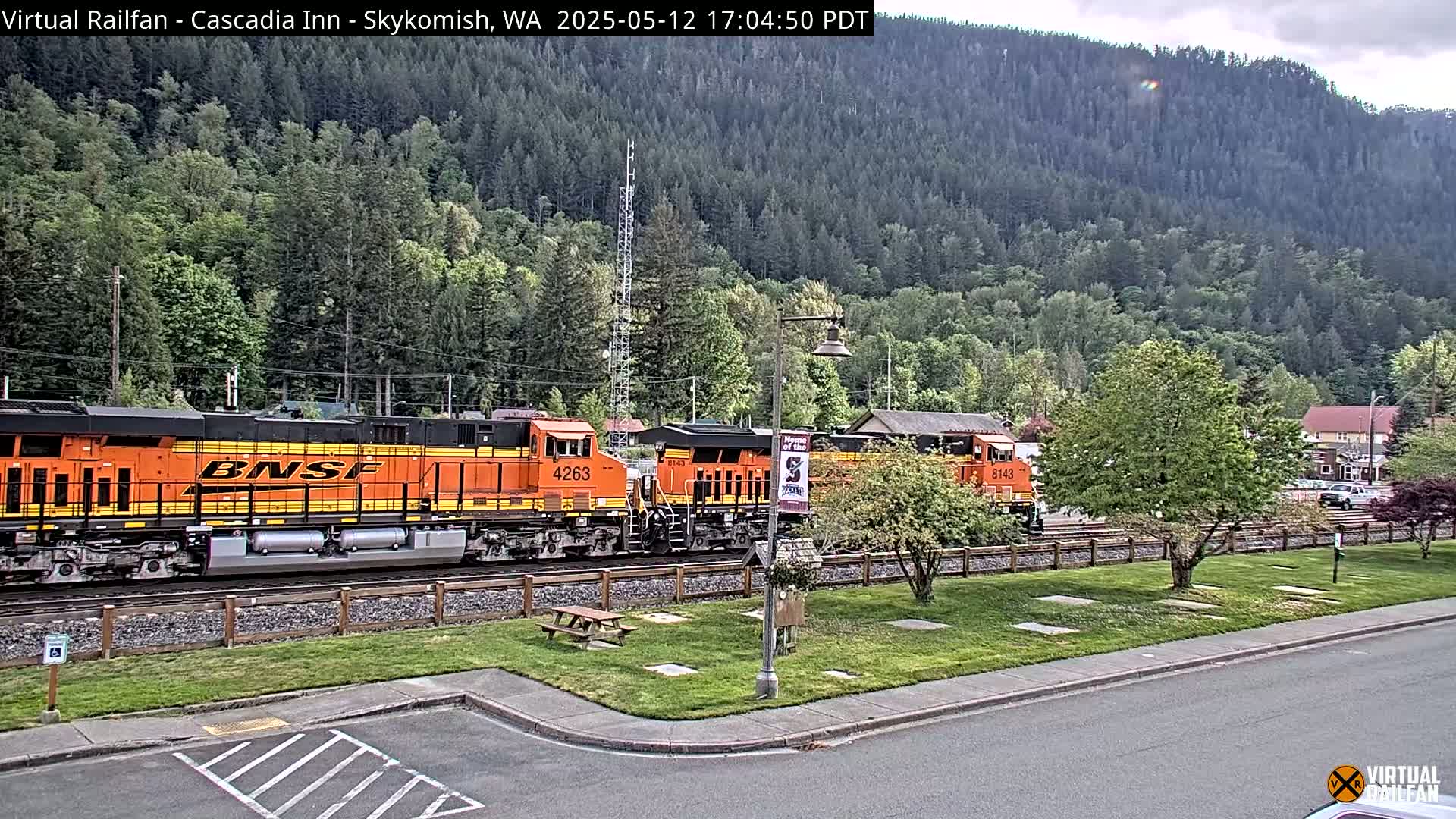 Two orange BNSF locomotives are parked on a train track next to a grassy area with trees and a hill covered in a forest in the background, under an overcast sky.