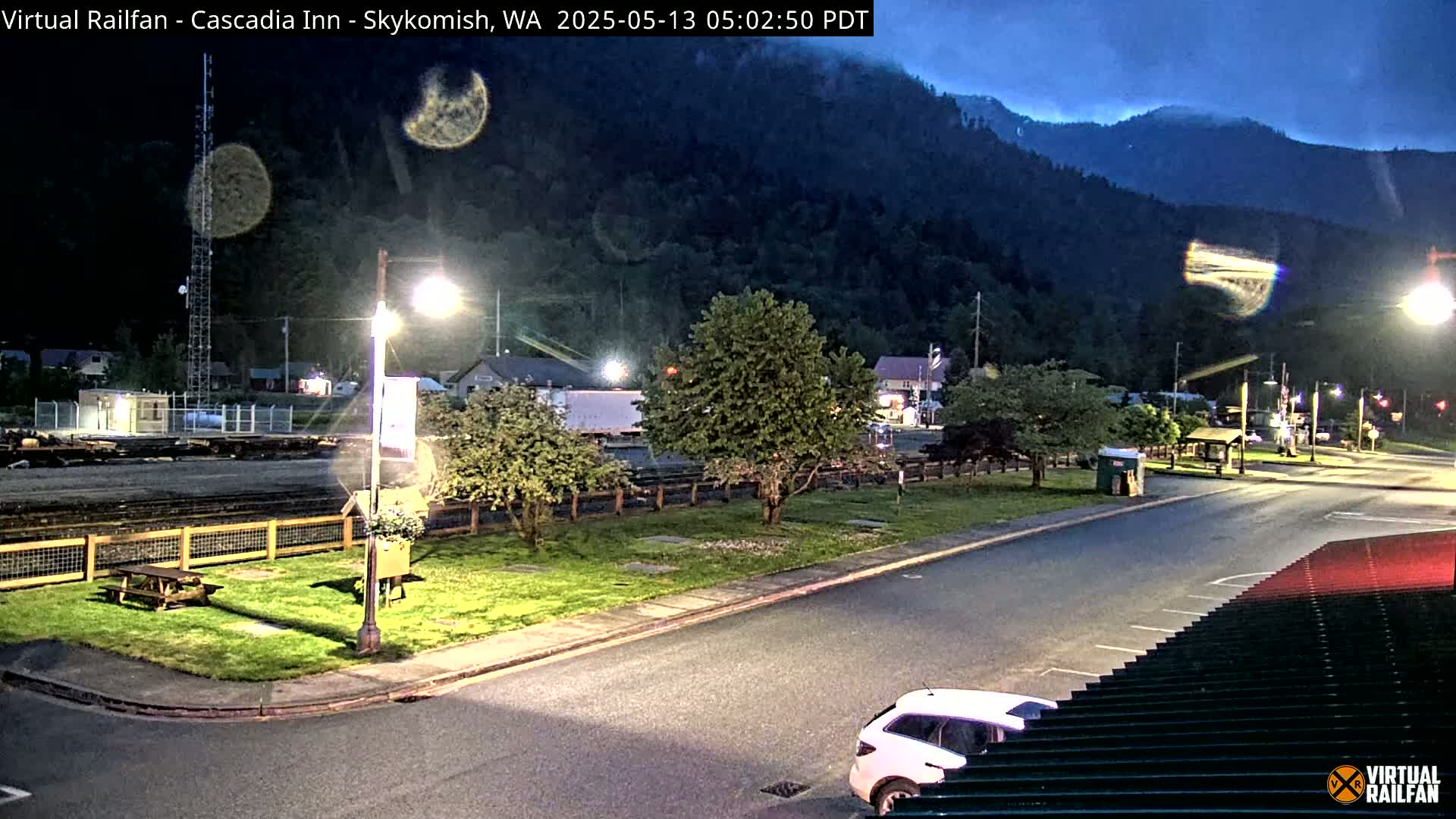 A nighttime view of a town beside a mountain, featuring a road, grassy areas, a picnic table, and a car, under a dark, possibly cloudy sky.