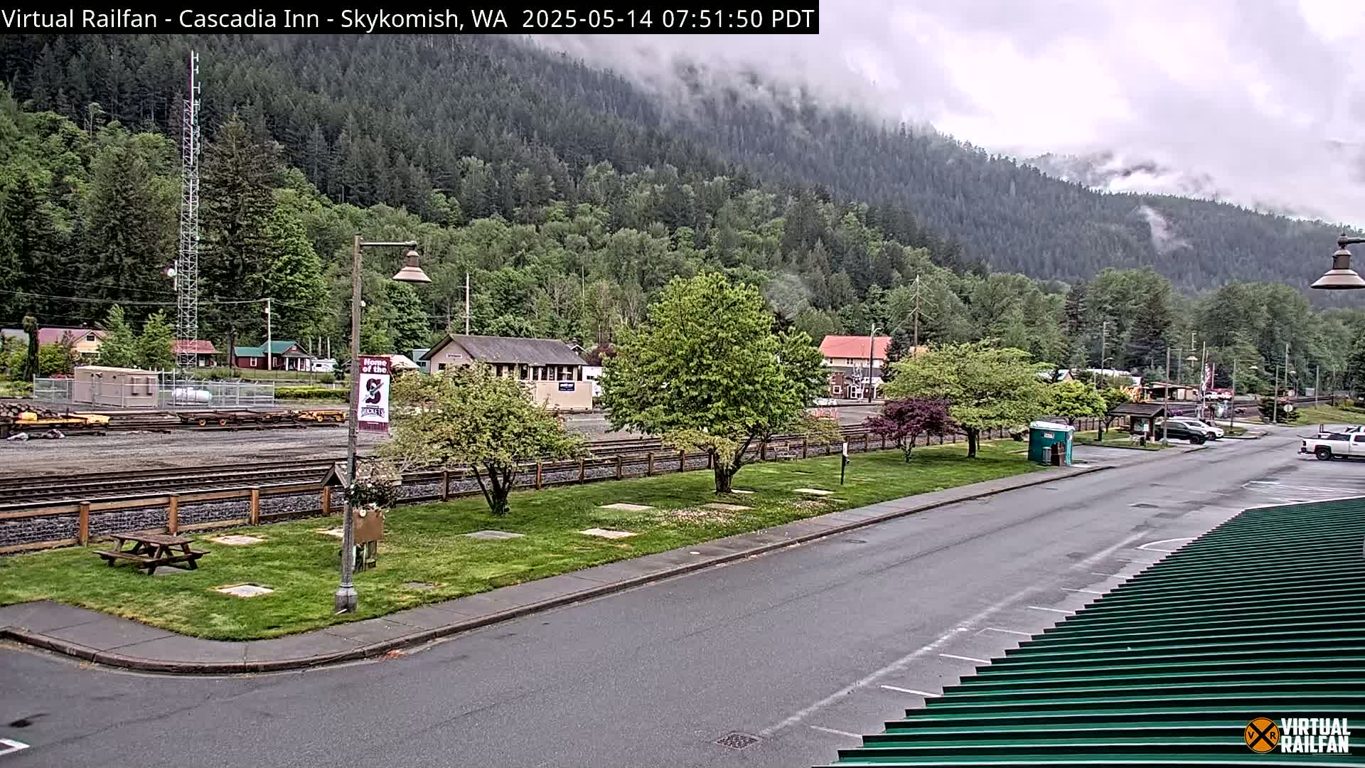A cloudy, overcast day reveals a view of a small town nestled in a valley, with train tracks and buildings visible, alongside a grassy area with trees and a picnic table.