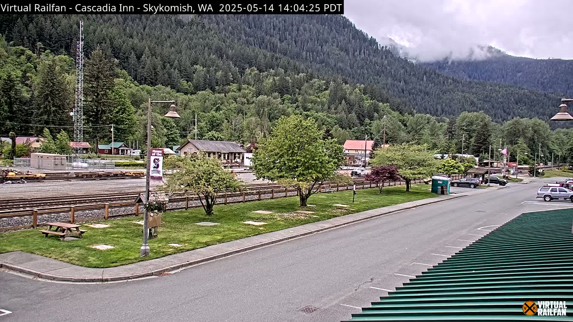 A small town nestled in a valley beneath a cloudy sky, features a train track, parked cars, and a grassy area with trees.