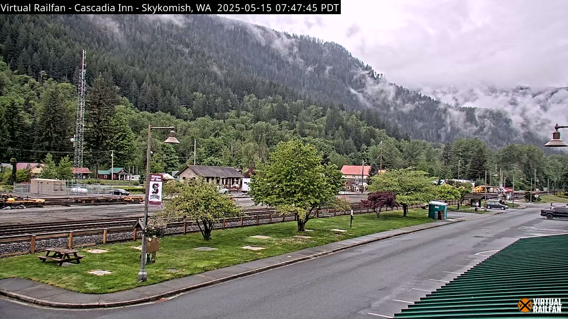 A cloudy day reveals a small town nestled at the base of a forested mountain shrouded in mist, with railroad tracks running through its center.