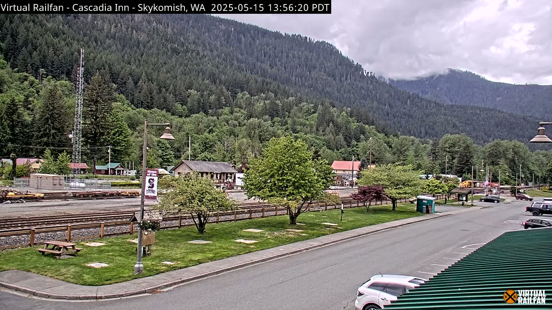 A partly cloudy day reveals a town nestled beside a mountain range, featuring train tracks, a few cars, and some small buildings.