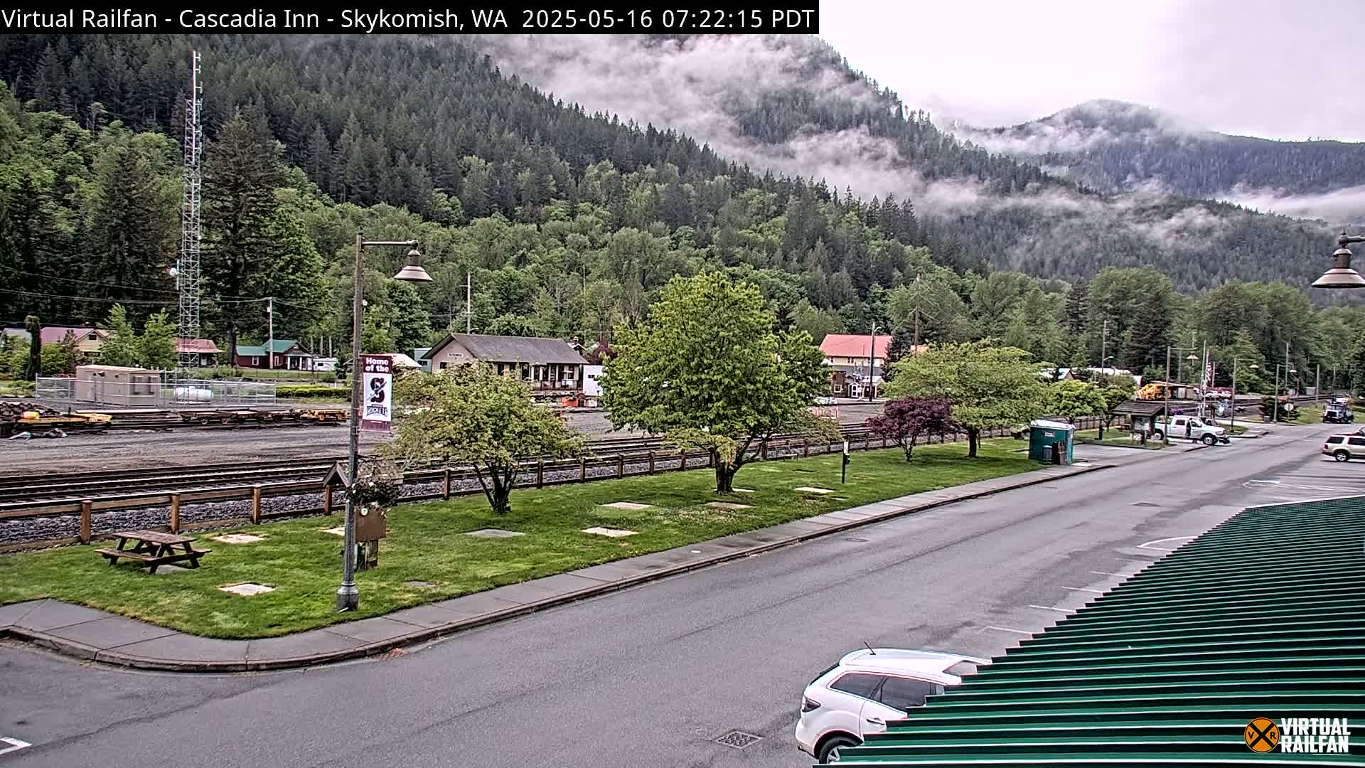 A misty mountainside overlooks a small town with a train depot, parking lot, and a few vehicles.