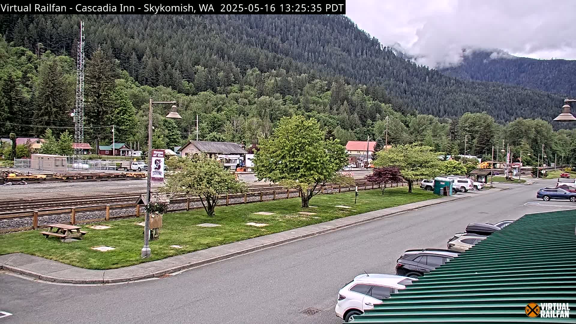A small town nestled in a valley under a cloudy sky, featuring a train track, a parking lot with several cars, and green landscaping.