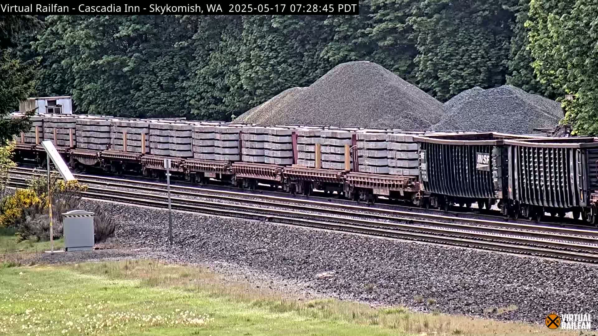 A long freight train carrying rectangular loads and followed by several empty gondolas is moving along a railroad track next to piles of gravel under a partly cloudy sky.