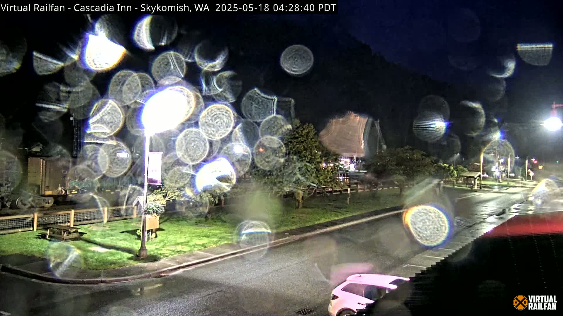 Rain-streaked nighttime view of a street scene with a pink car, streetlights, and a grassy area near a rail line, all blurred by rain droplets on a window.