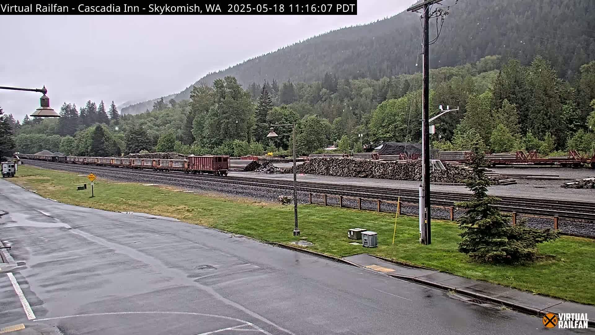 A long freight train carrying logs is visible near a lumber yard in a lightly raining landscape, with a forested hillside in the background.