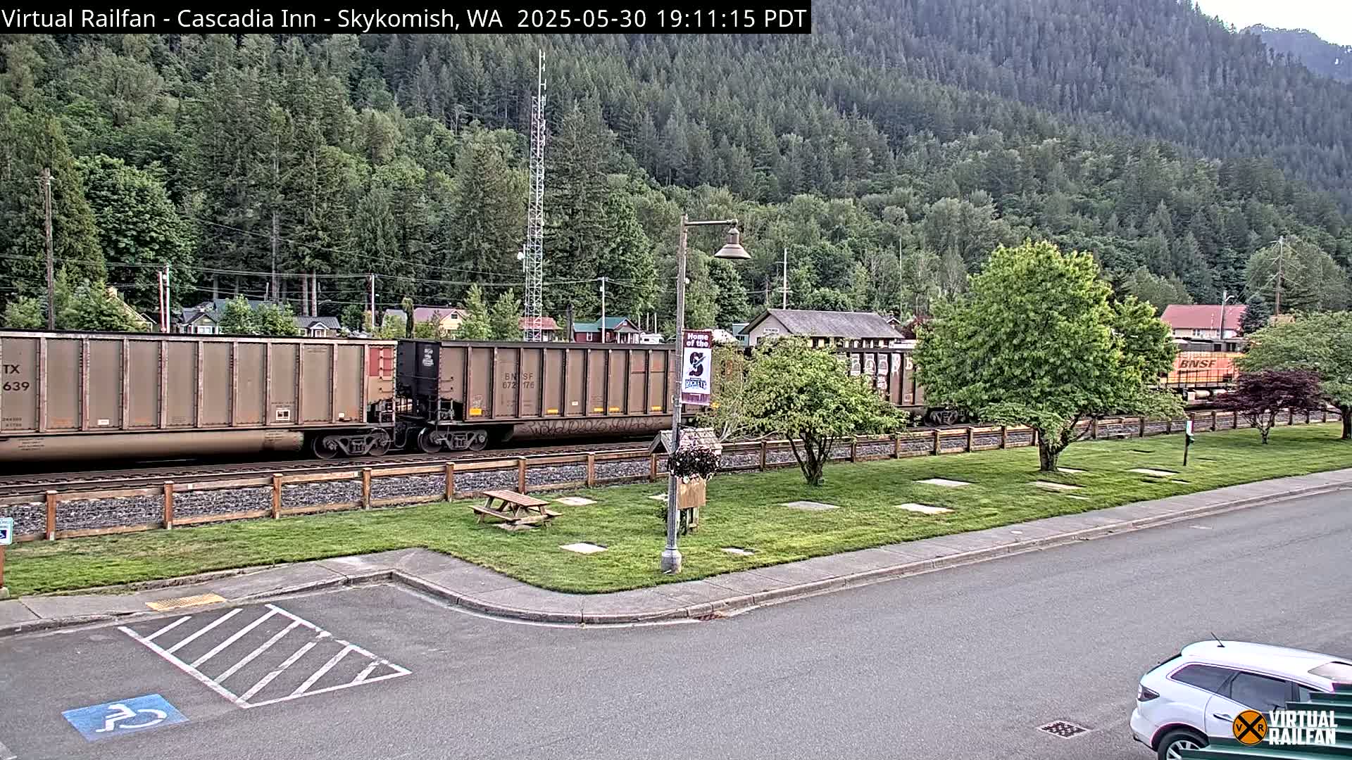 A freight train passes by a grassy area with a picnic table and a small parking lot, in front of a forested hillside on a partly cloudy day.