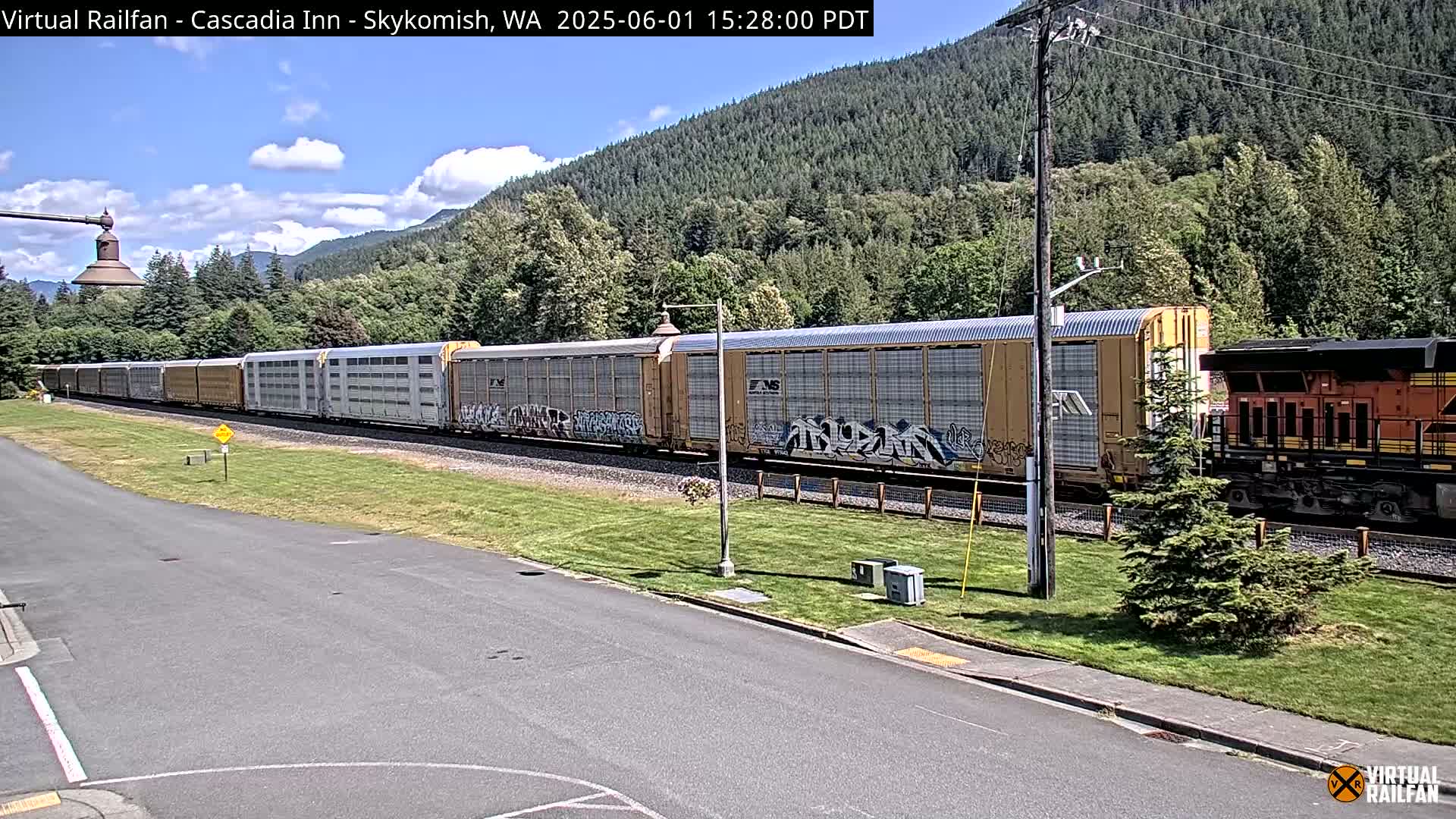 A long freight train passes by on a sunny day, alongside a road and a grassy area next to a forested hillside.