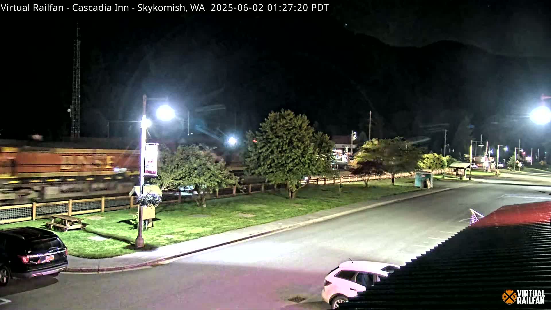 A long freight train passes by at night, near a parking lot with cars and some landscaping, under streetlights.