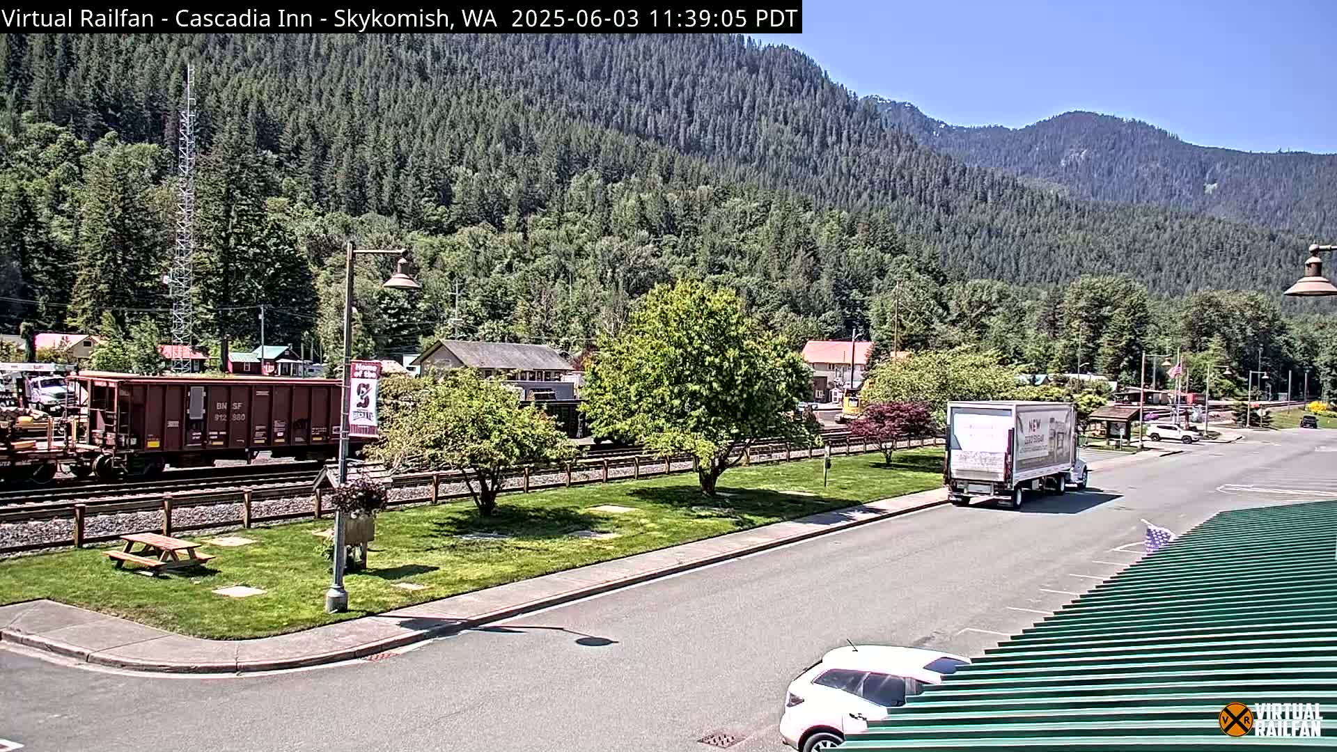 A sunny day in a town nestled in a valley surrounded by lush green mountains, with a train and some vehicles visible near a paved area.