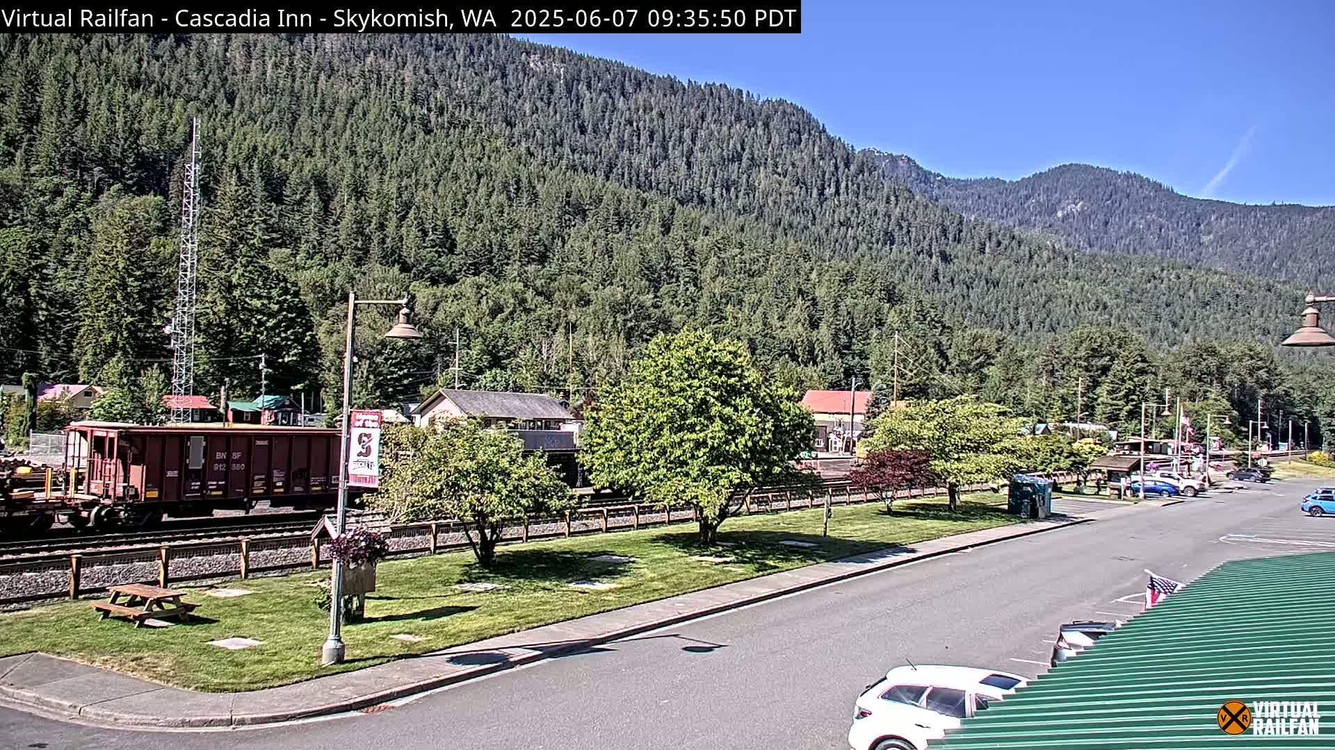A railroad car sits on a track beside a road in a town nestled in a valley, with a sunny sky and lush green mountains in the background.