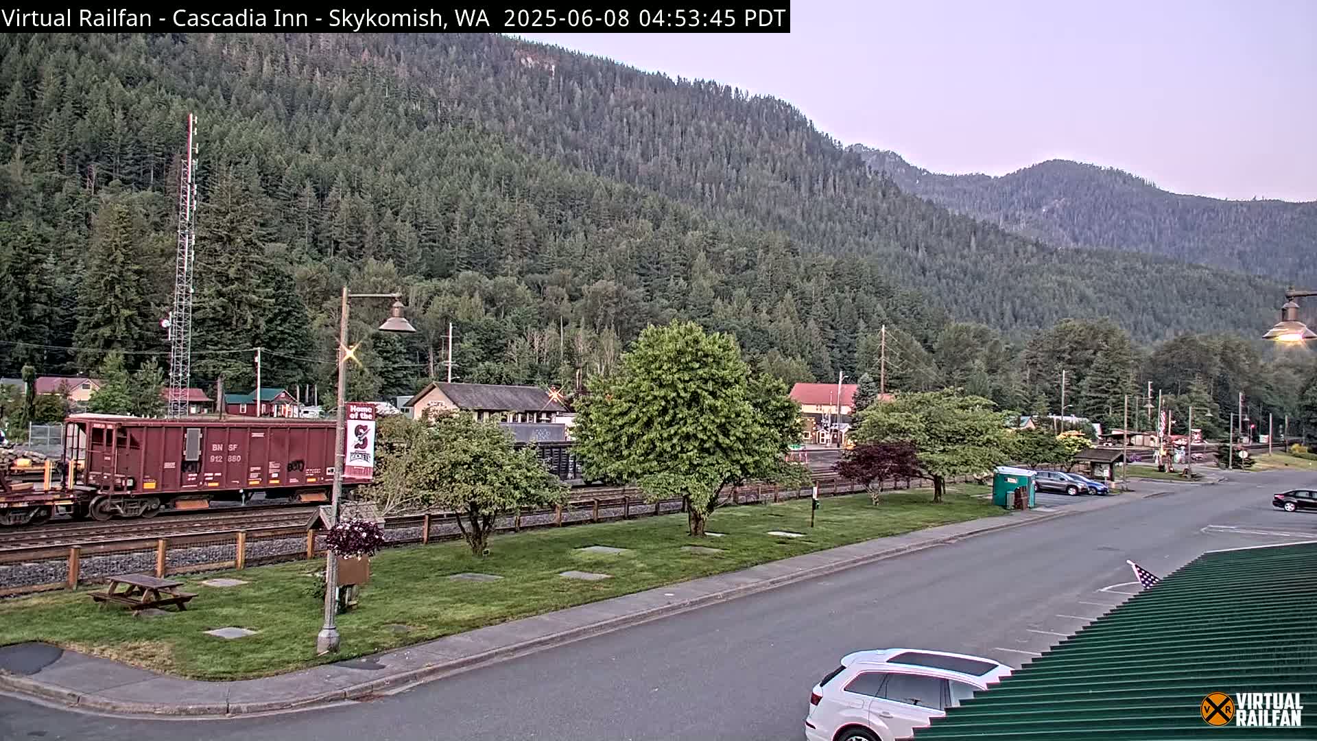 A railroad car sits on a track next to a road in a small town nestled against a lush green mountainside under a twilight sky.