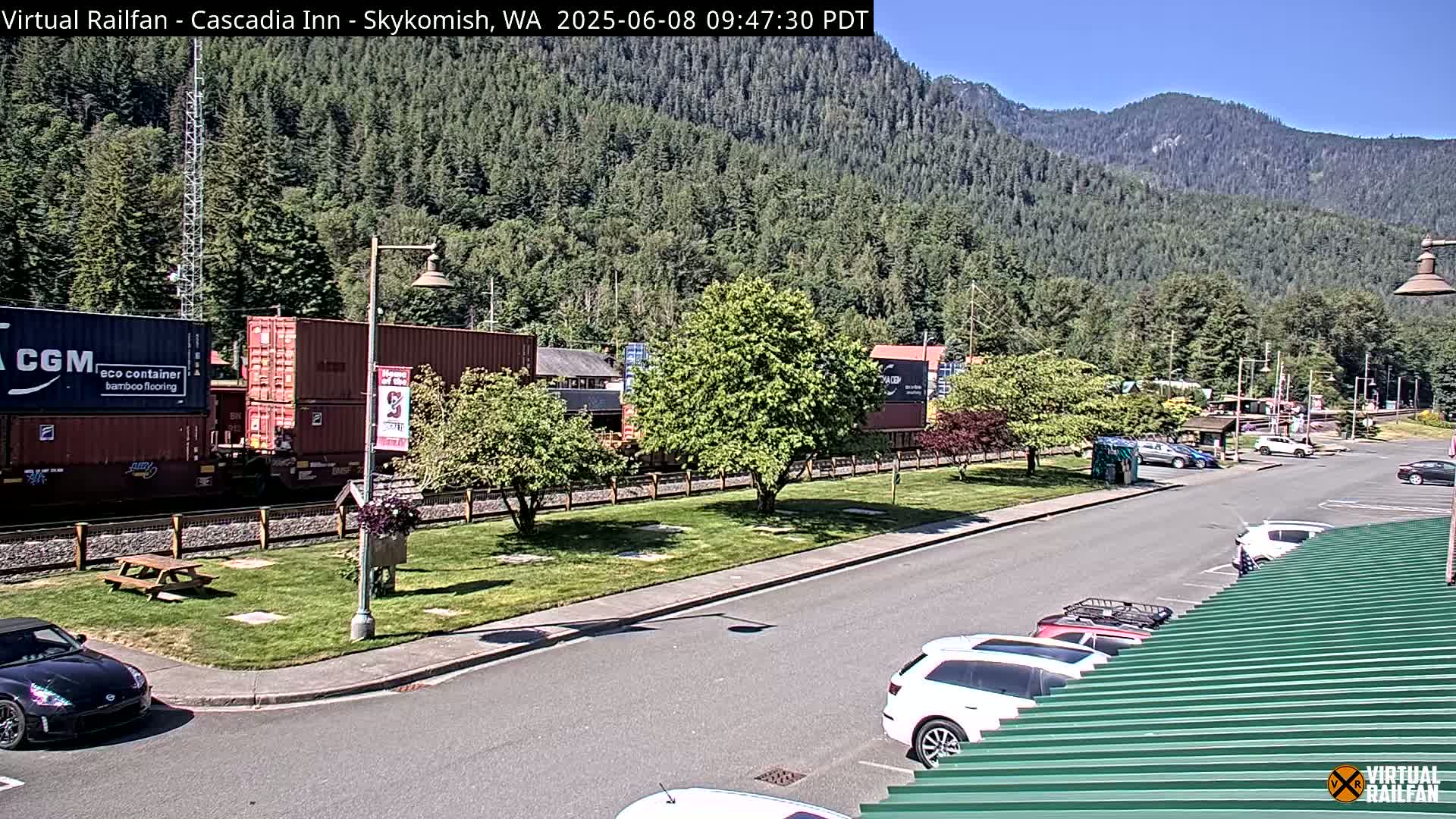 A sunny day reveals a train with shipping containers parked beside a roadside area with cars and a grassy lawn, all set against a backdrop of a mountain covered in evergreen trees.