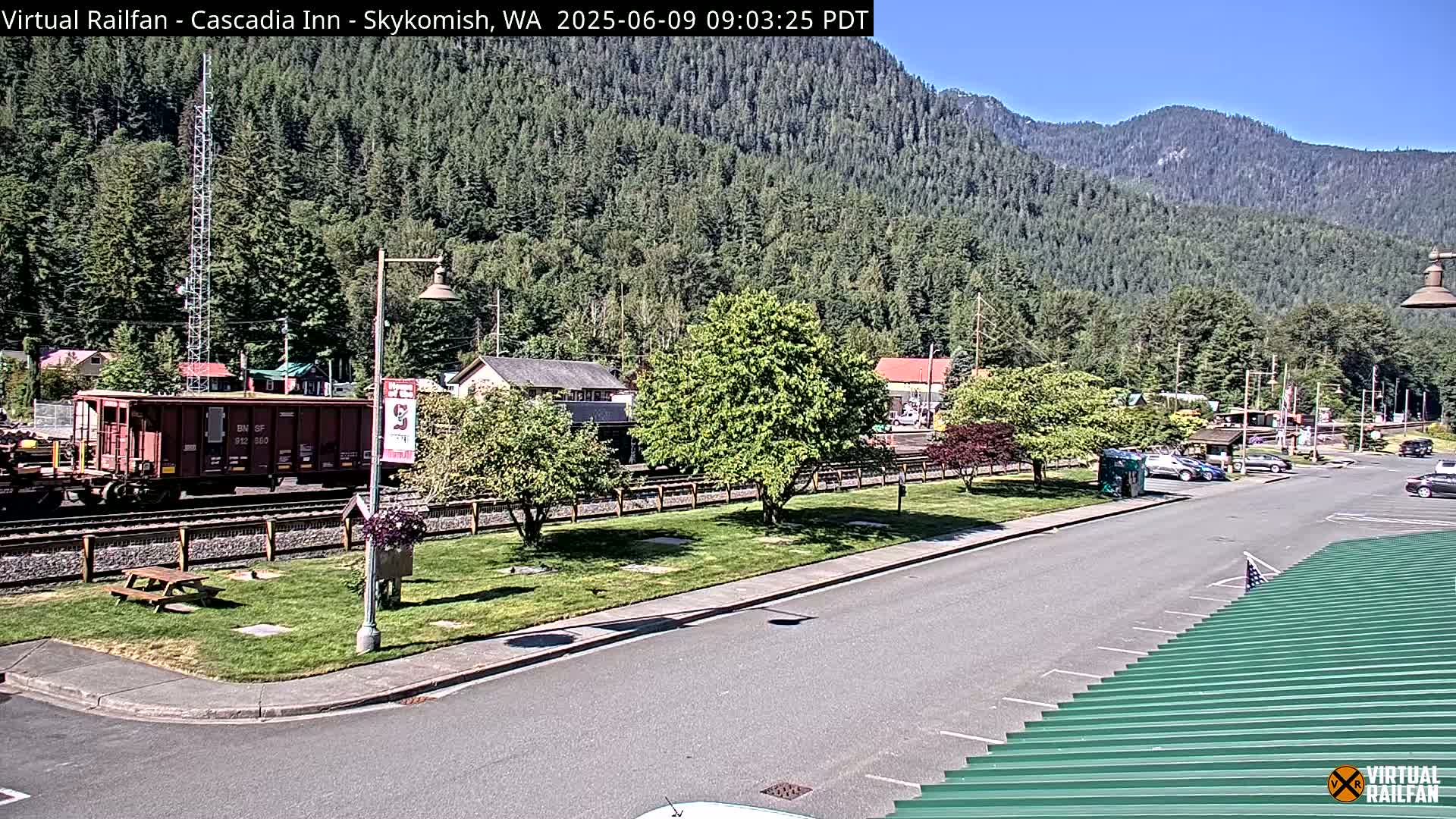 A freight train is parked near a small town nestled in a valley surrounded by lush green mountains under a sunny sky.
