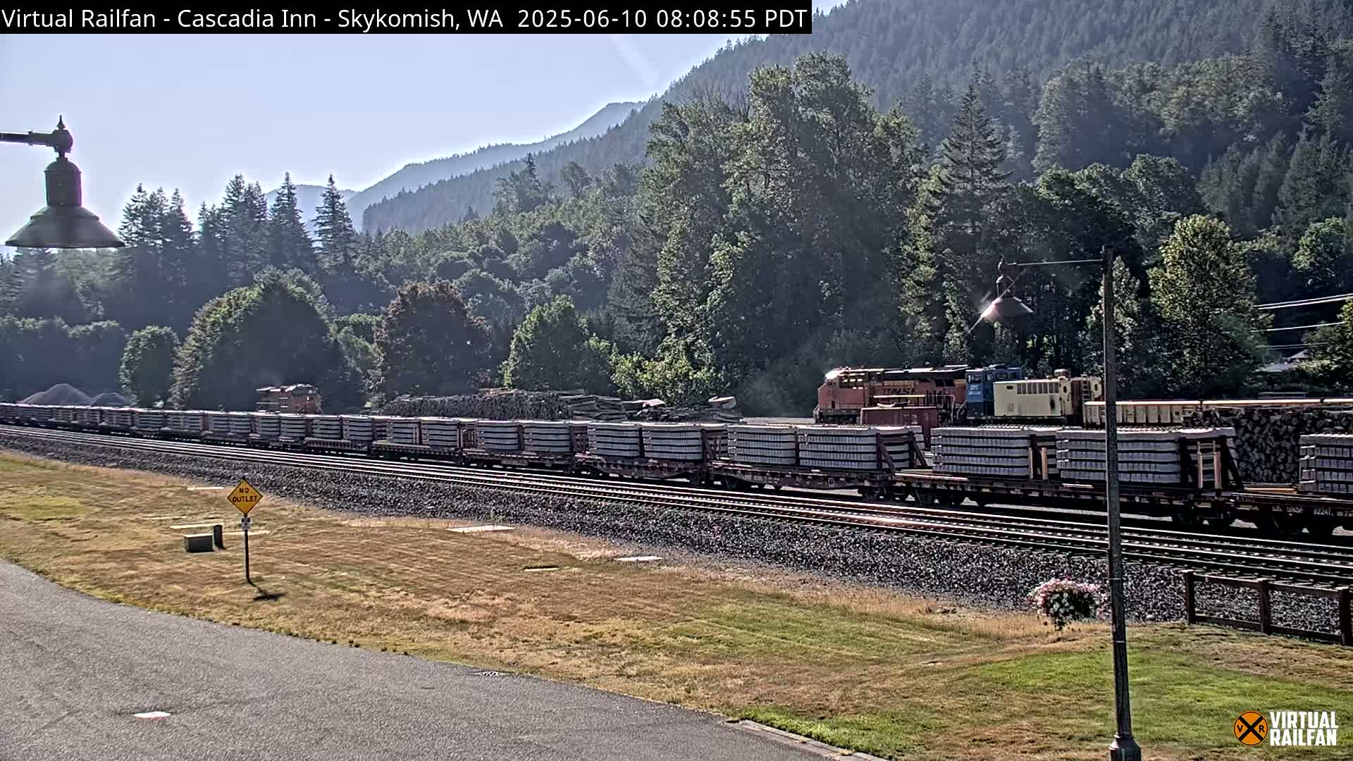A long freight train carrying stacked materials is visible on a railroad track, set against a backdrop of lush green forested hills on a sunny day.