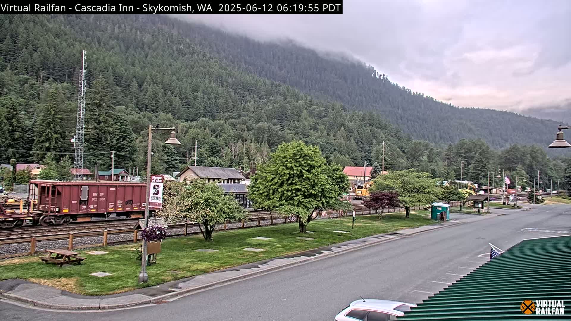 A railroad car sits on tracks beside a street in a small town nestled against a mountainside under an overcast sky.
