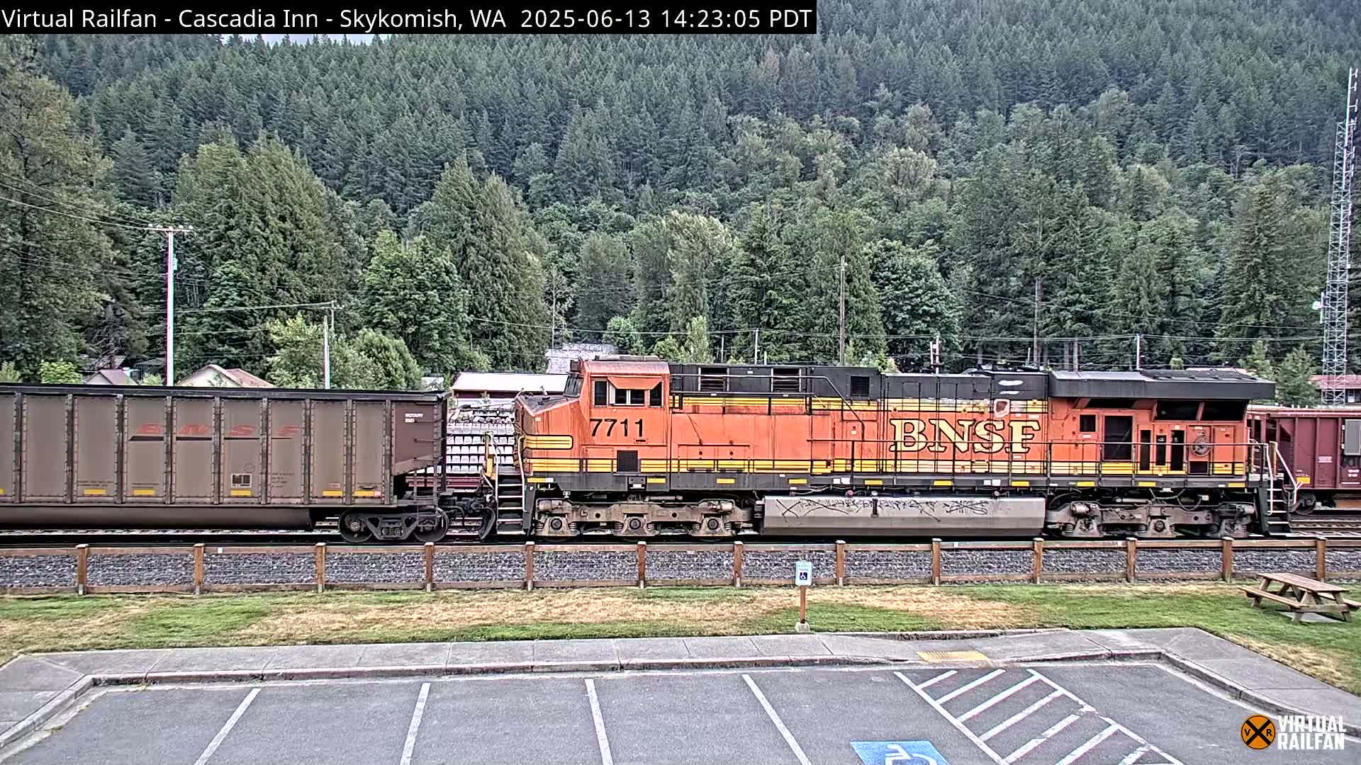 An orange BNSF freight train, pulled by a locomotive numbered 7711, travels past a grassy area and parking lot against a backdrop of a lush green forest.