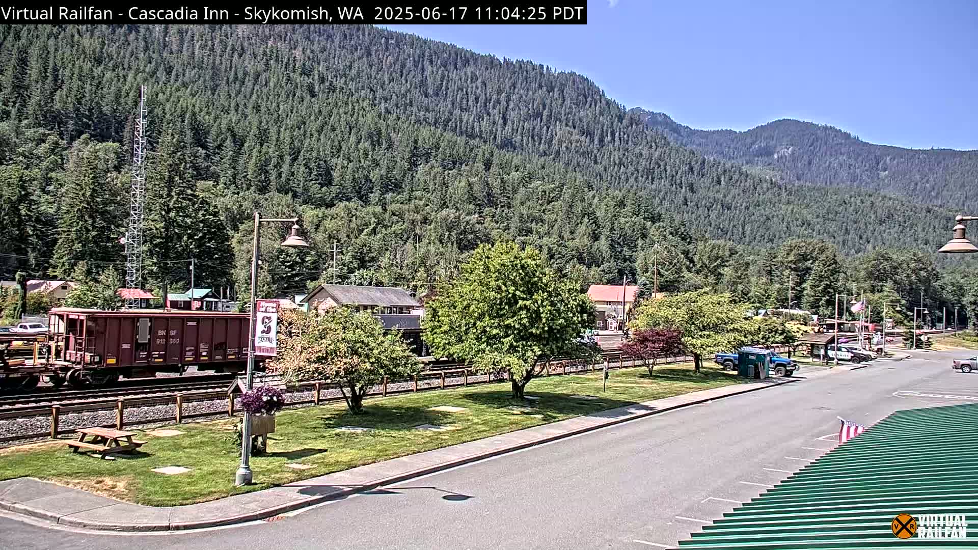 A train car sits on railroad tracks next to a small town nestled in a valley beneath a mountain covered in evergreen trees on a sunny day.