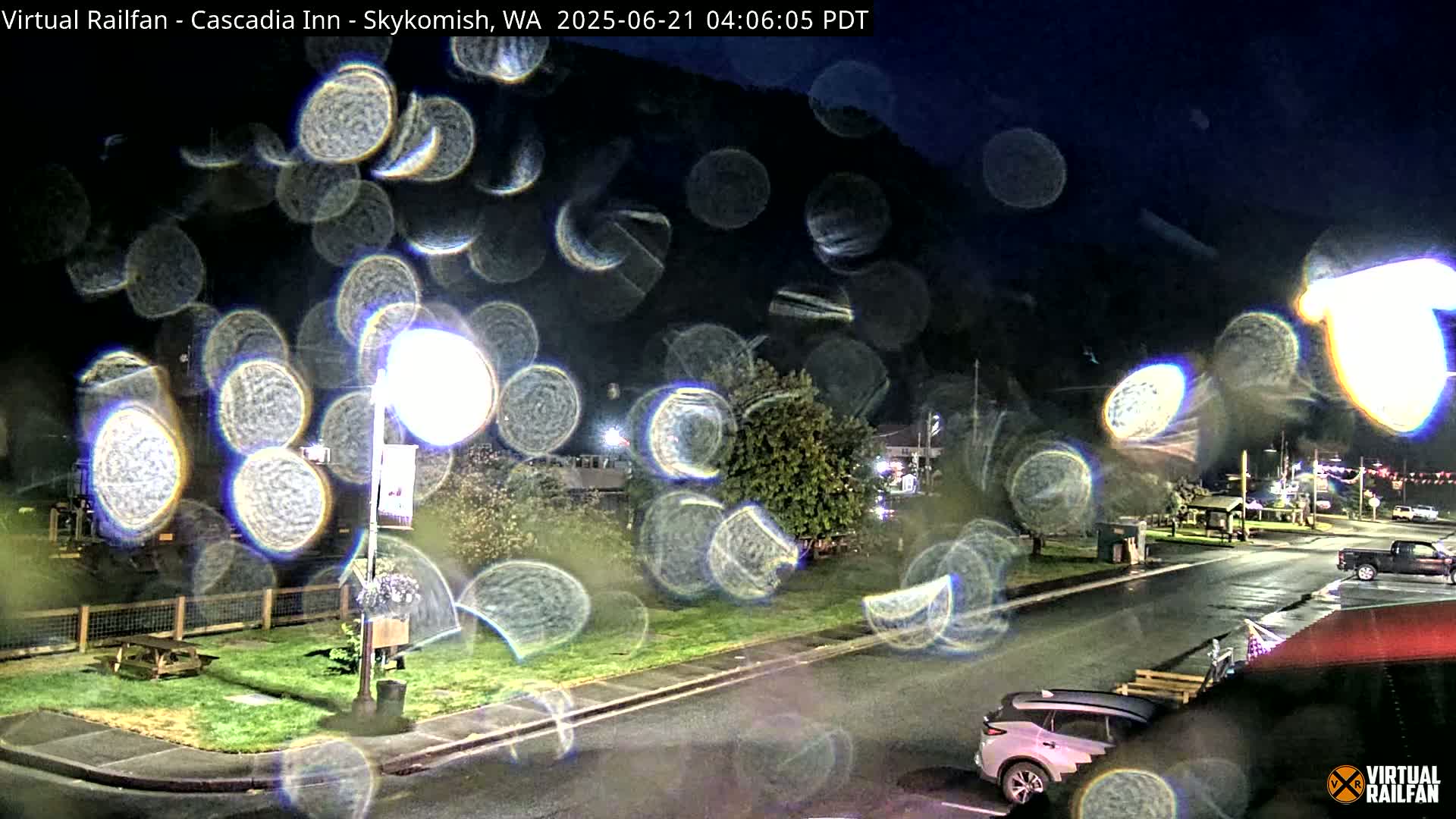 A nighttime view of a street scene, obscured by rain droplets on a window, shows a parking lot, cars, and a grassy area with a lamppost.