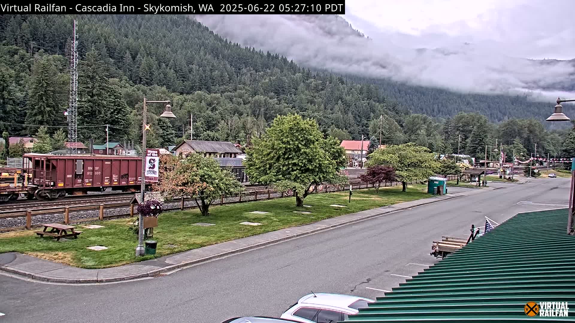 A railroad car sits on the tracks beside a town nestled in a valley partially obscured by fog, under an overcast sky.