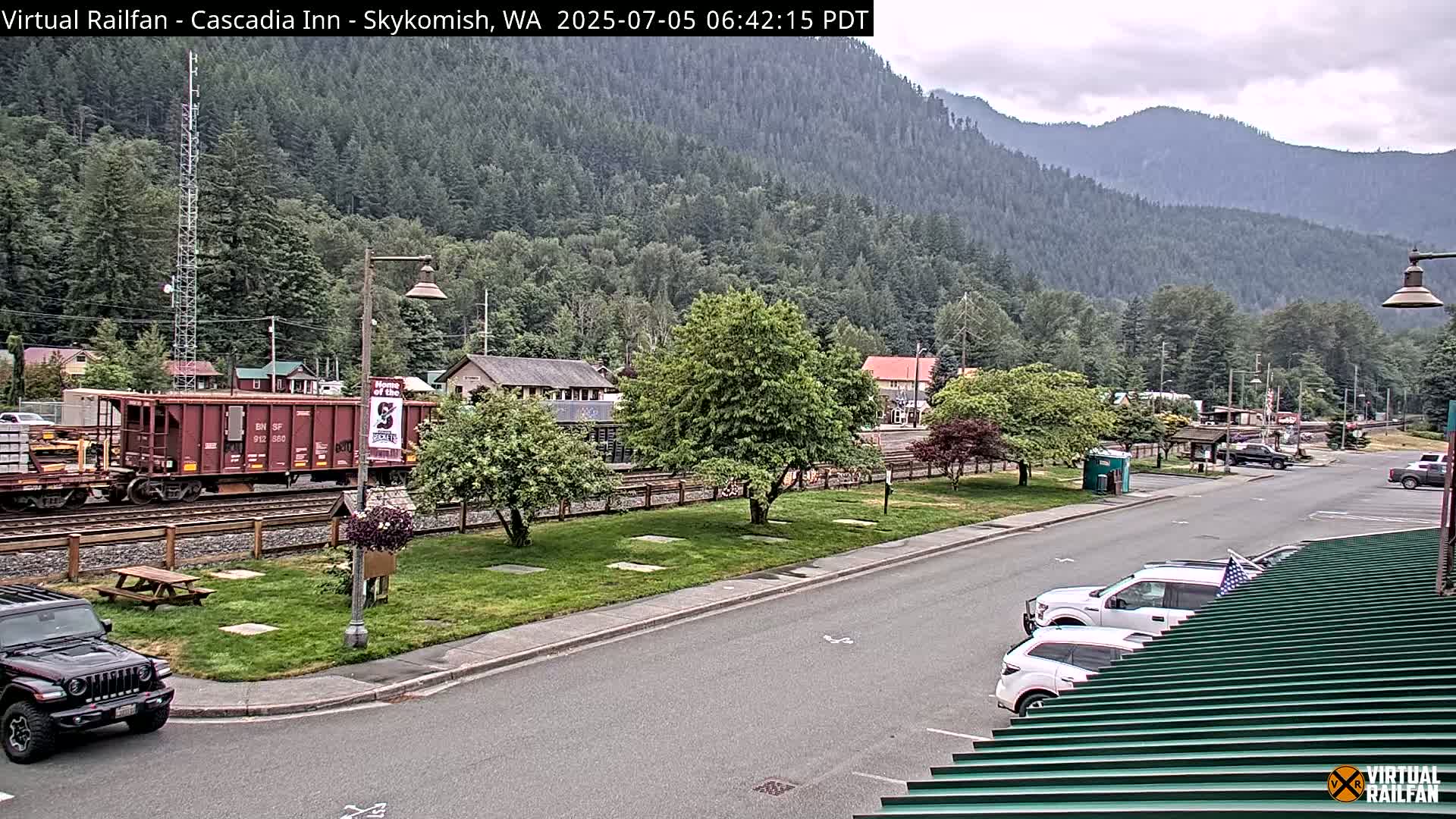A railroad car sits on the tracks beside a small town nestled in a valley between mountains under an overcast sky.