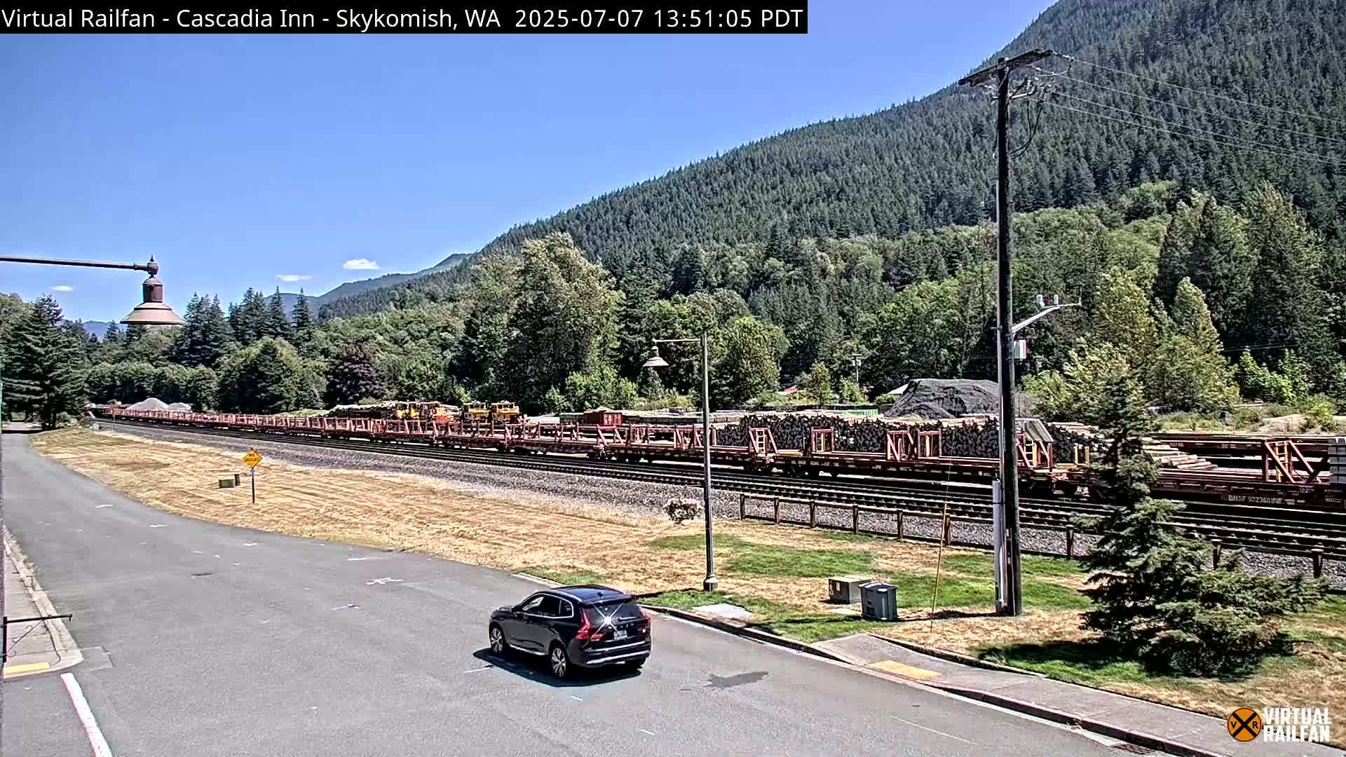 A long freight train carrying logs is parked on a railroad track next to a road, with a dark-colored SUV parked nearby, all under a clear, sunny sky and surrounded by lush green mountains.