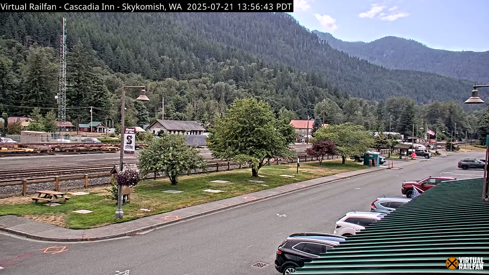 A partly sunny view of a town nestled in a valley surrounded by mountains, featuring a parking lot with several cars and a railway line with freight cars in the background.