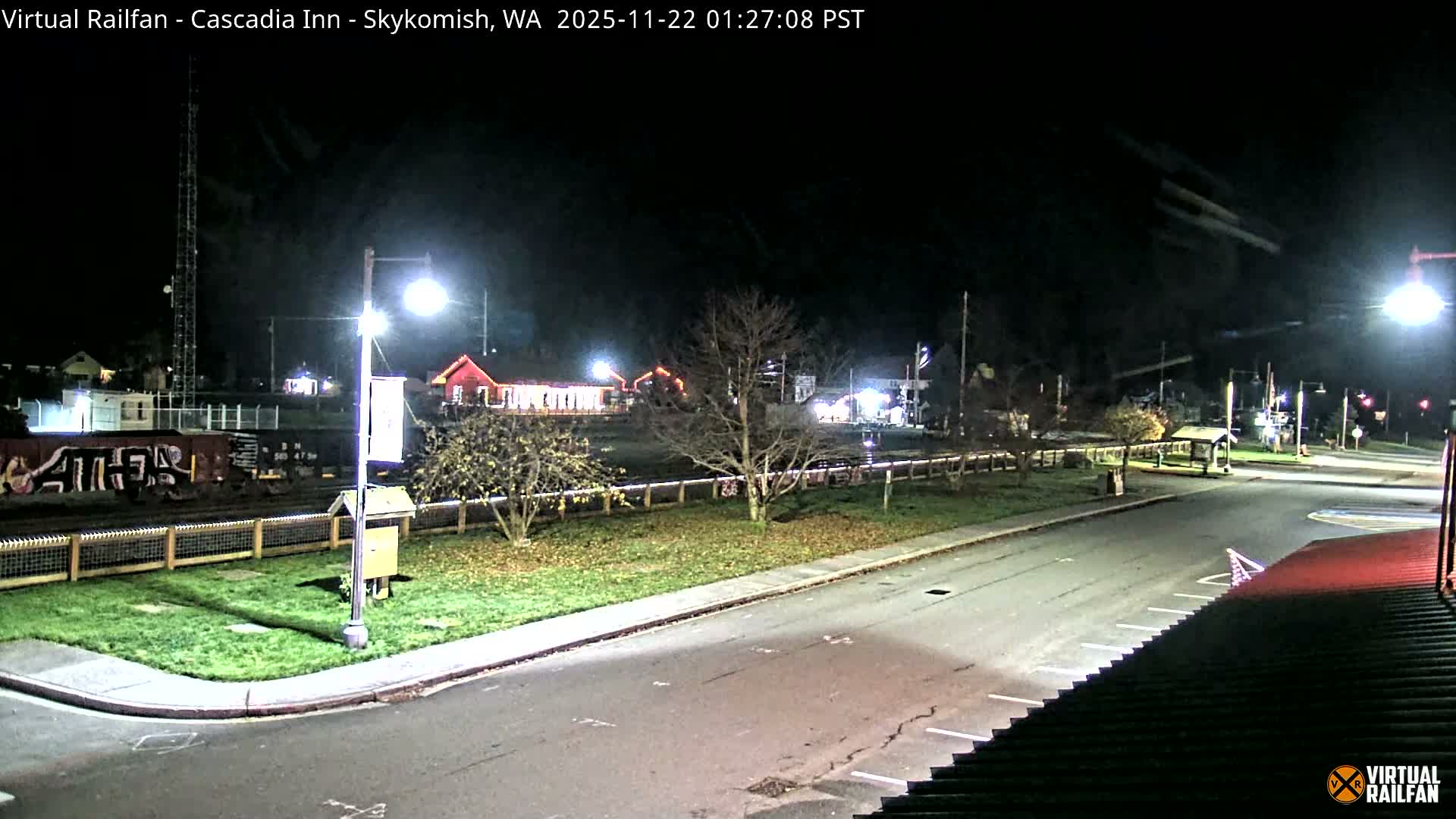 A clear night view of a small town reveals illuminated streets and buildings, a train car on tracks, and bare trees under the dark sky.