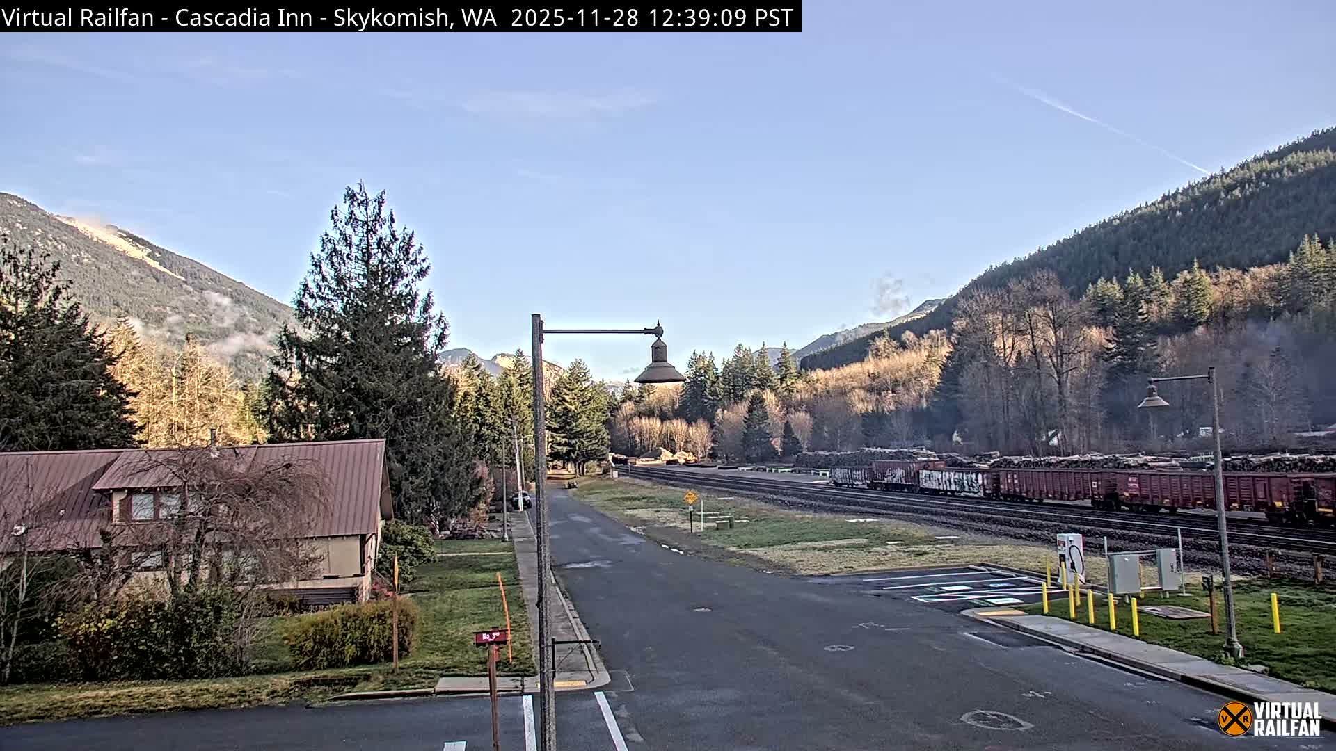 A long freight train carrying logs sits on tracks next to a road and a house, with densely forested mountains rising in the background under a clear blue sky.