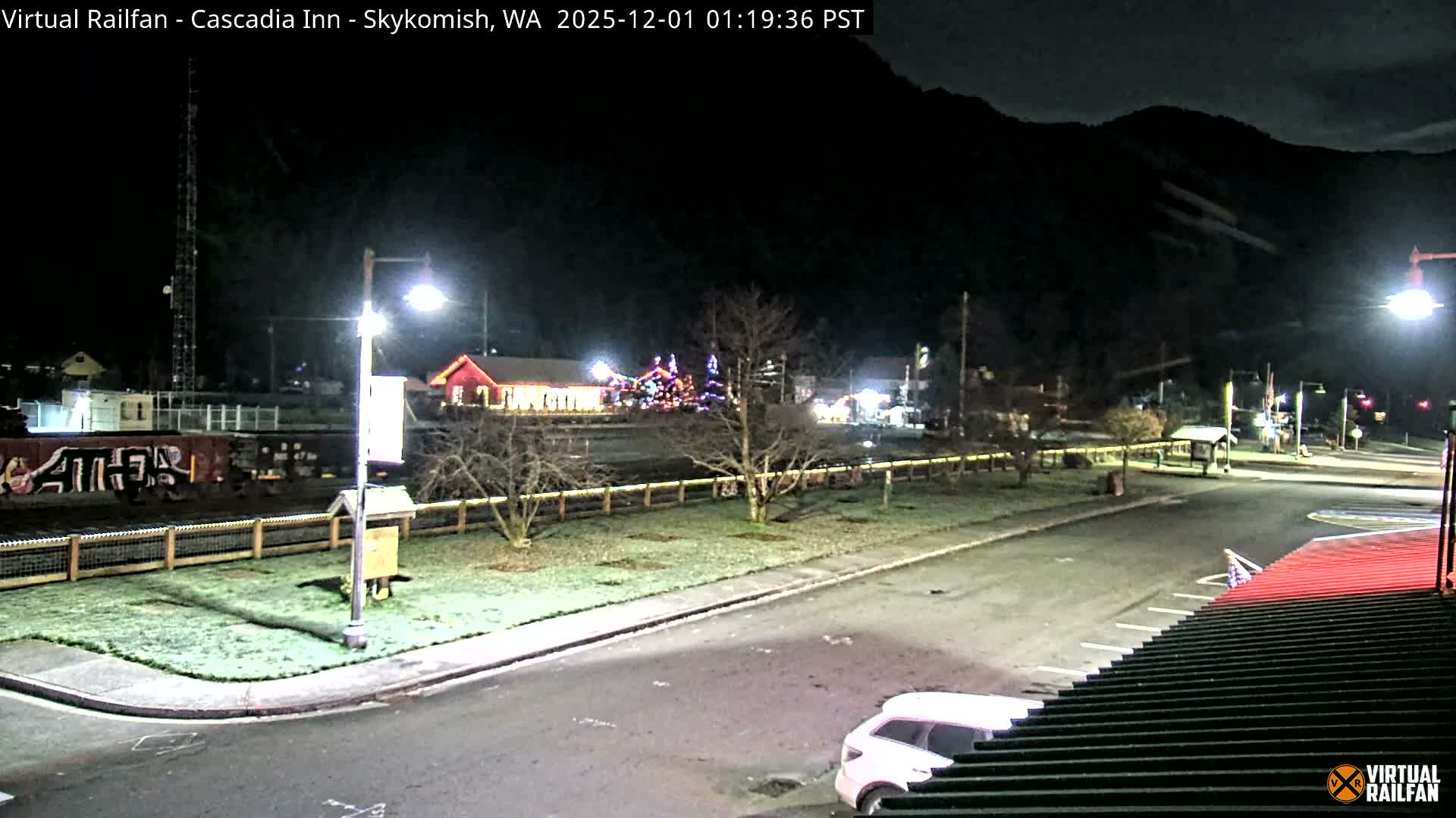 A quiet, illuminated small town street and park area are visible at night, bordered by train tracks with freight cars and buildings, one of which is adorned with colorful holiday lights, all under a clear, dark sky.