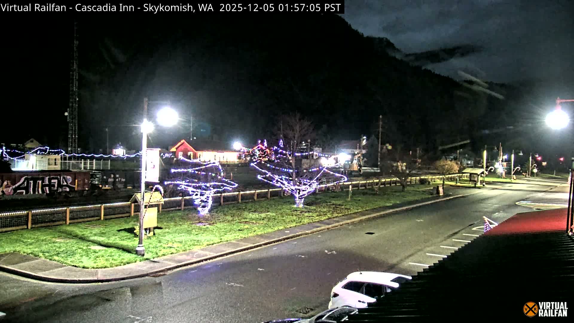 A wet night scene in a small town features a street and grassy median adorned with bare trees wrapped in festive string lights, a railroad track with a parked train car, and dark, cloud-covered mountains in the background.
