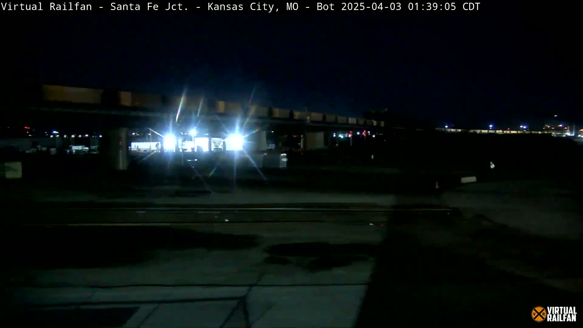 A train with bright headlights is seen at night approaching a railway station under a clear, dark sky.