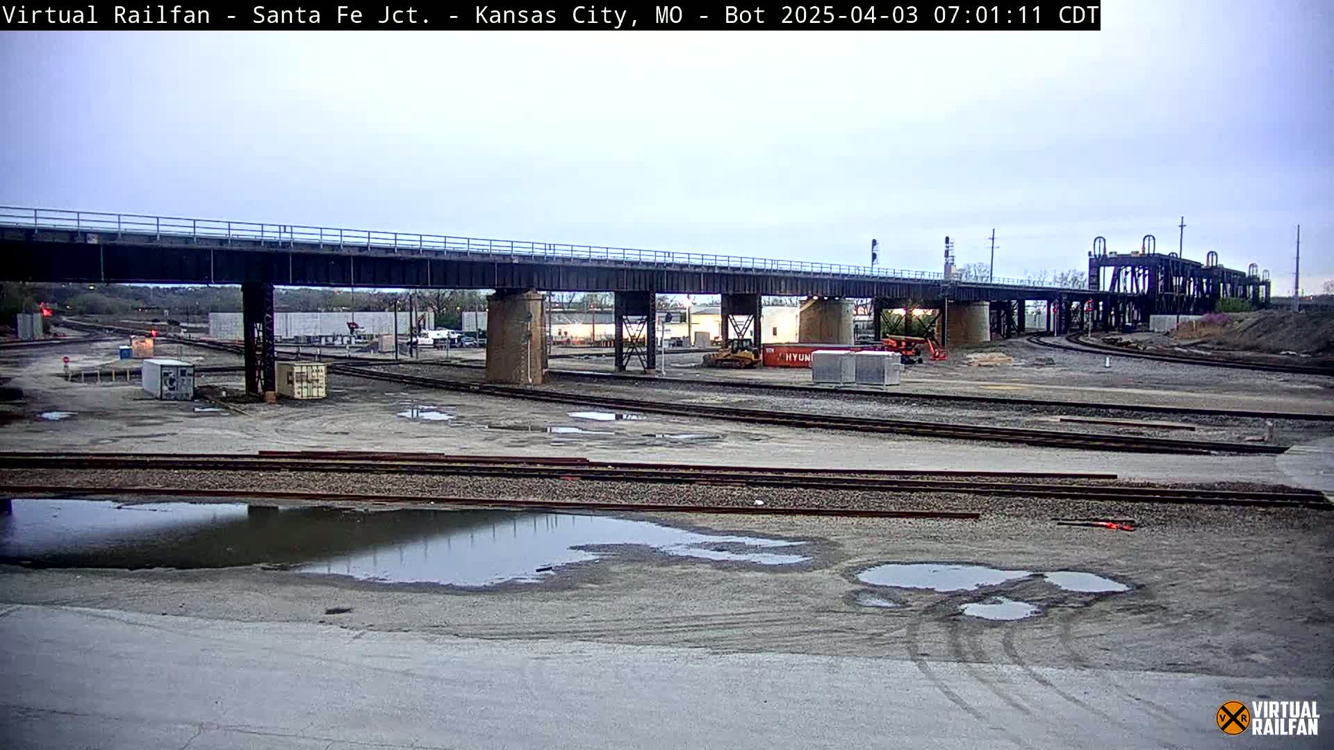 A railway yard with a large bridge overhead, under an overcast sky with some puddles on the ground.