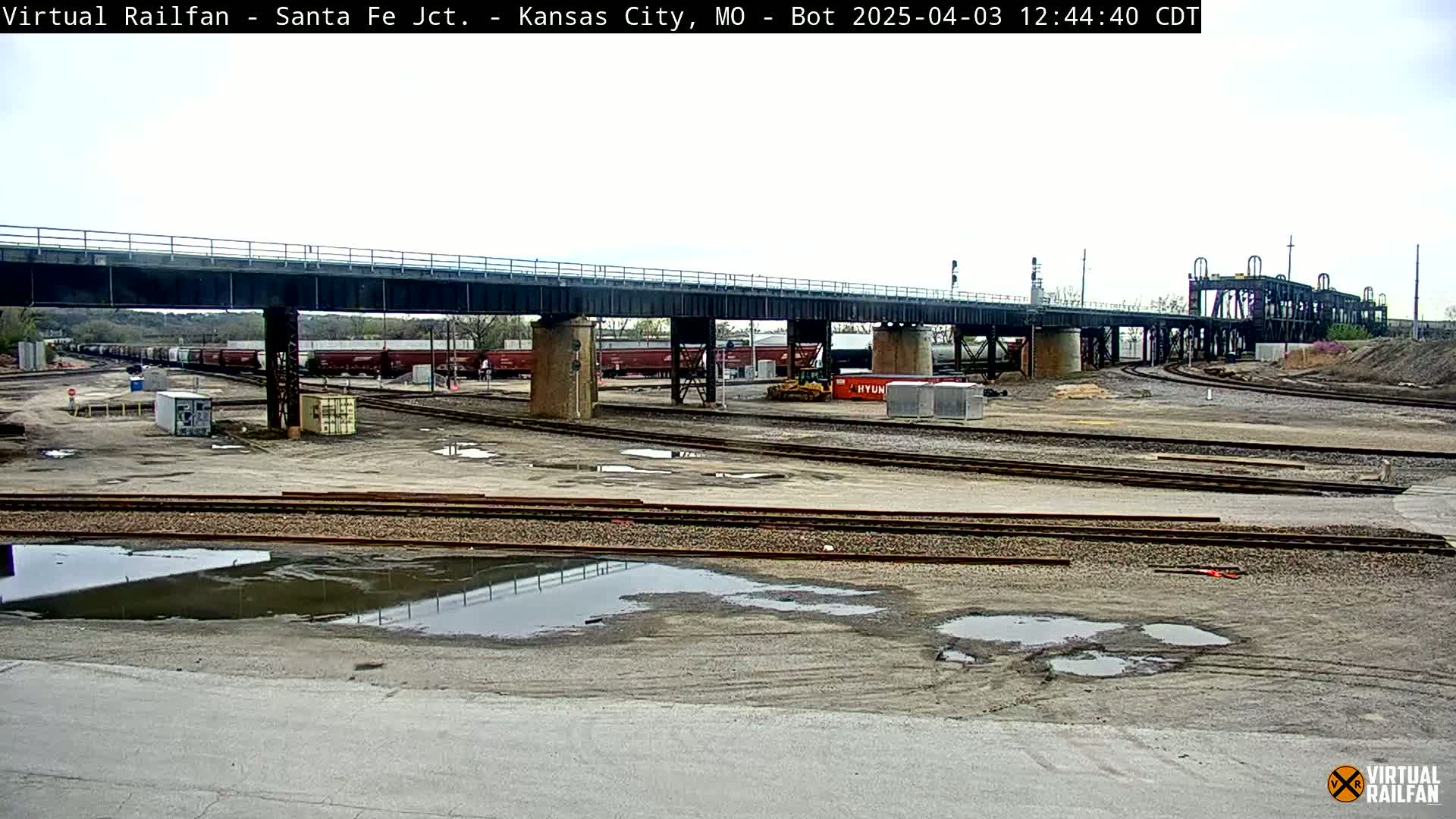 A railway yard with several train cars under a bridge, some puddles of water on the ground, and overcast skies.