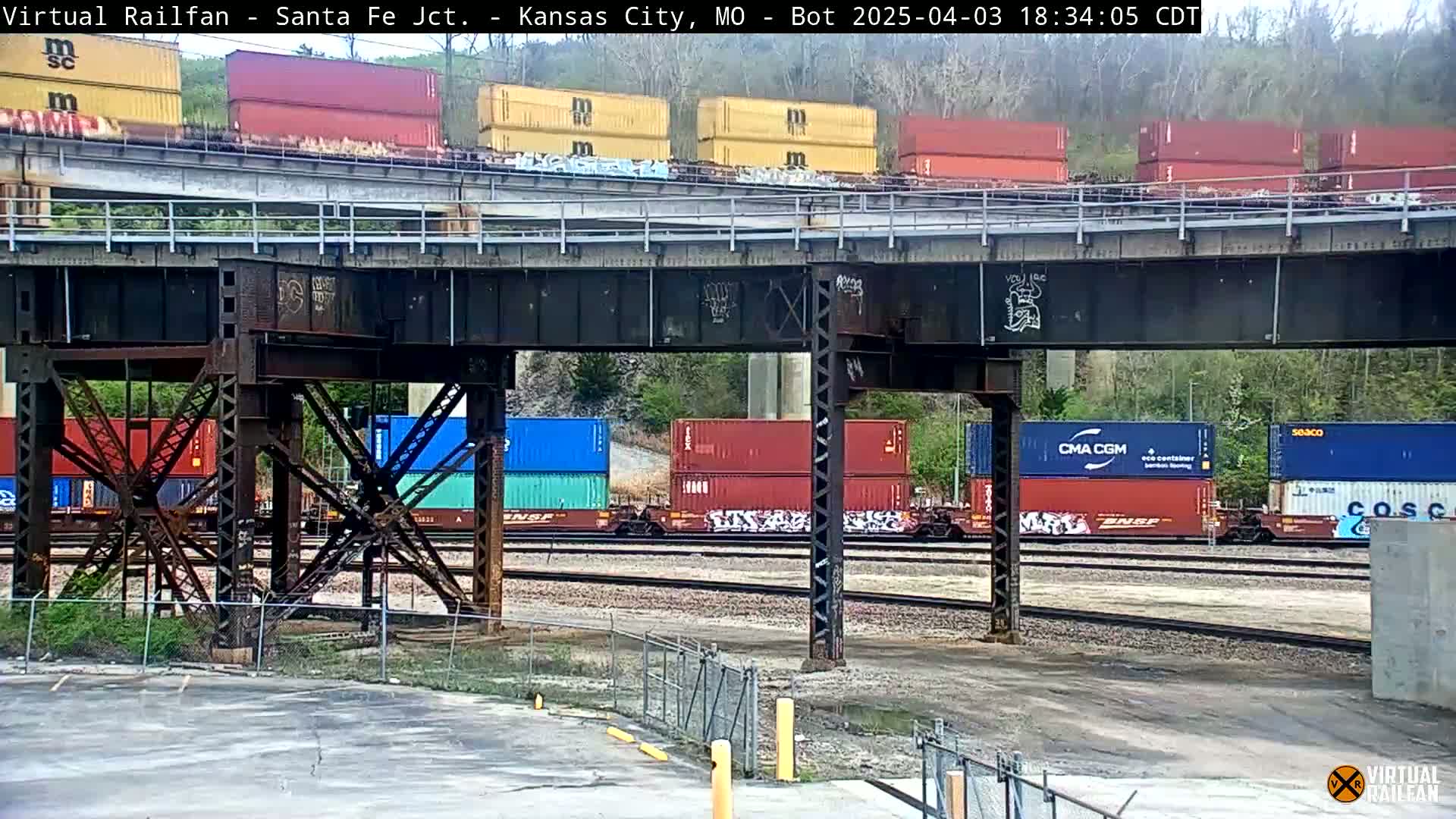 A train carrying numerous colorful shipping containers passes under a bridge on a partly cloudy day.