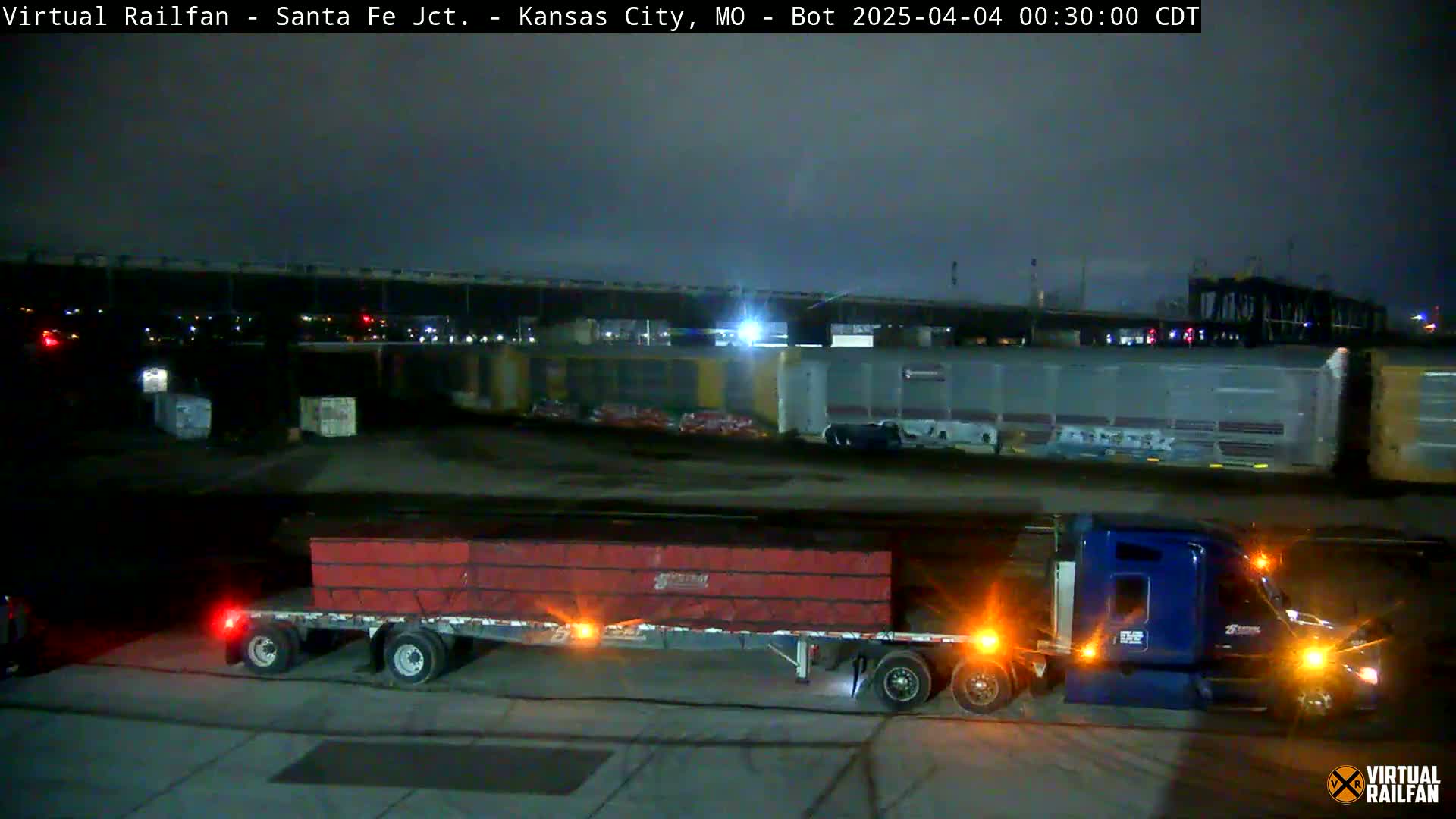 A blue semi-truck hauling a flatbed trailer loaded with reddish-brown cargo is parked at night near a train in a dimly lit industrial area.