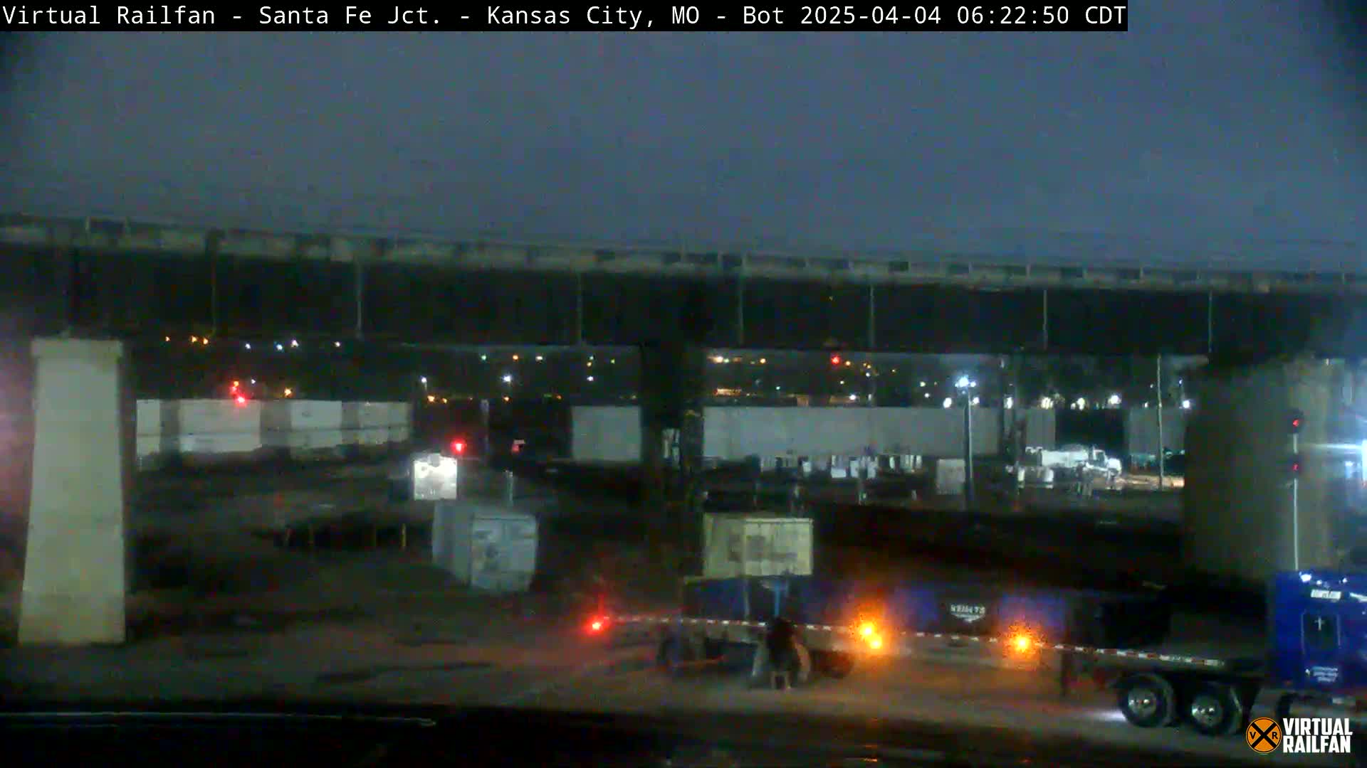 Under a dark night sky, a nighttime view shows several large trucks and shipping containers beneath a highway overpass.