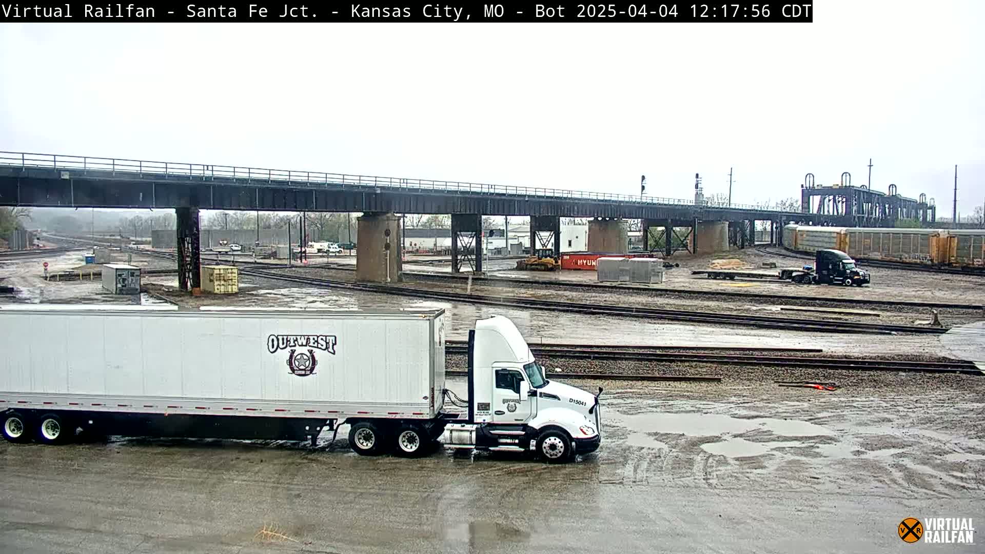 A large white semi-truck is parked in a rail yard under an overcast sky, with puddles on the wet ground and several railway tracks visible.