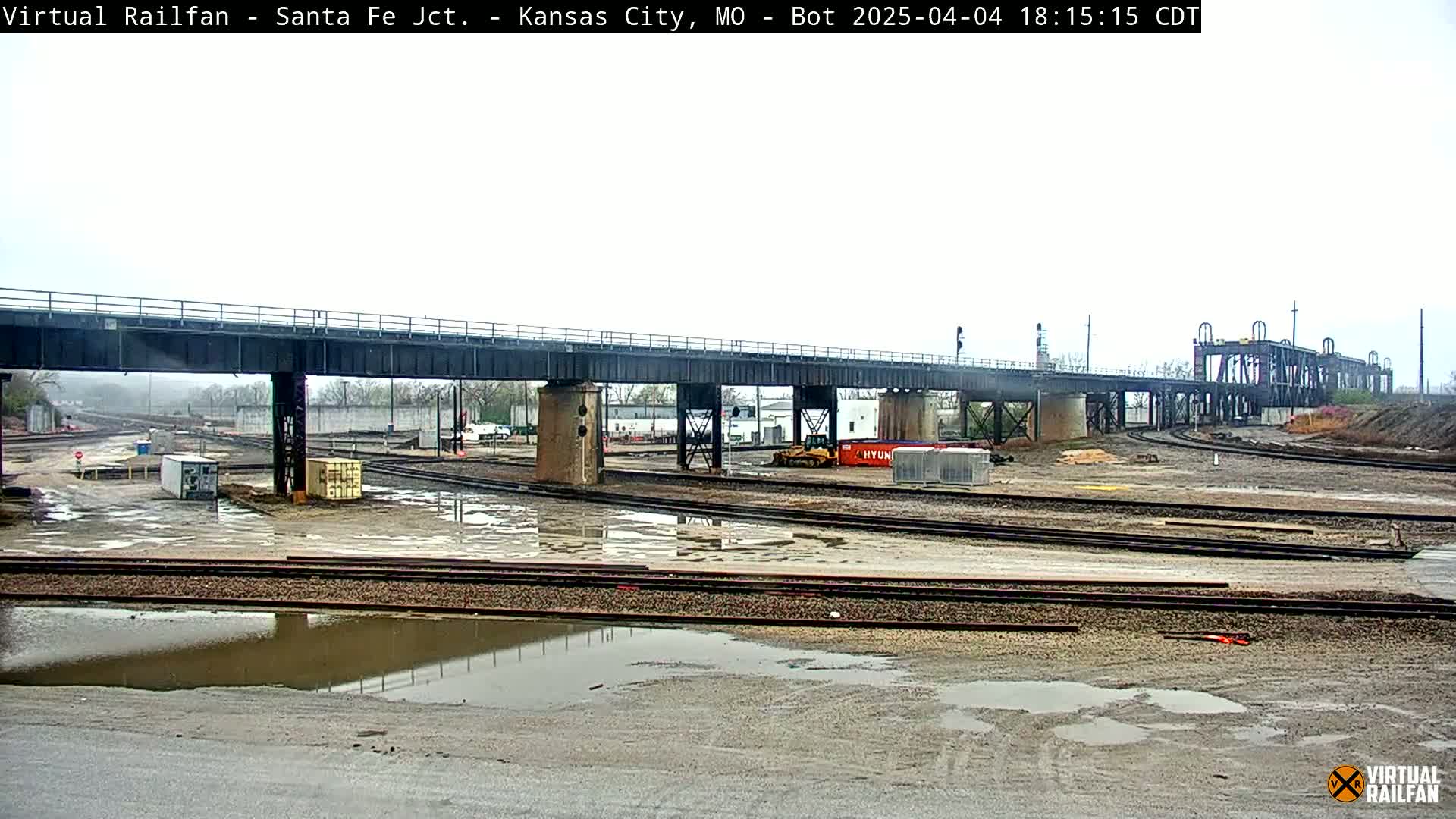 A railway yard with a bridge overhead is shown under overcast skies; puddles of water are present on the ground.