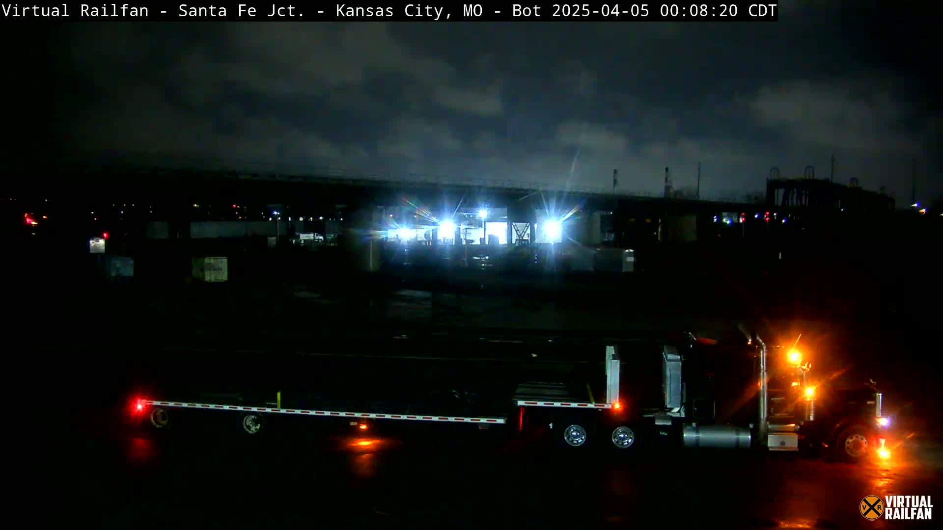 Under a cloudy night sky, a semi-truck with a flatbed trailer sits in a dimly lit industrial area.