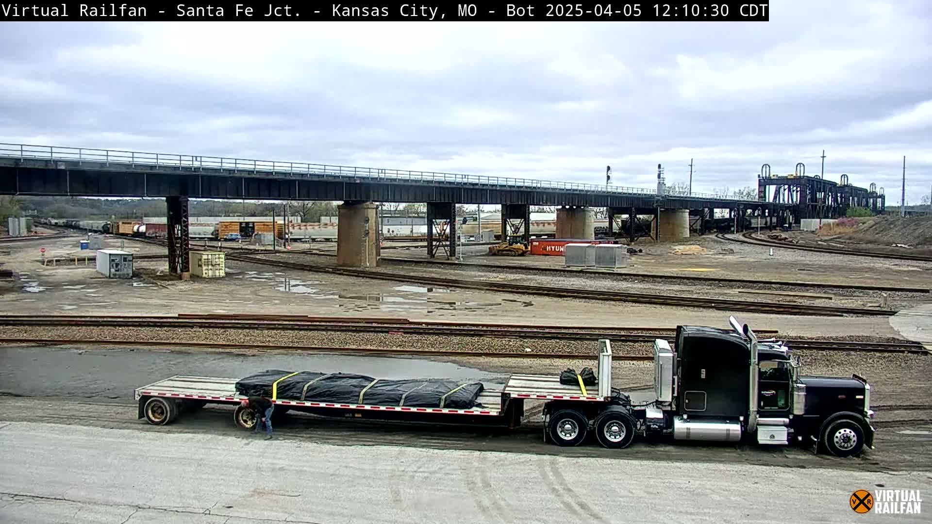 A black semi-truck hauling a flatbed trailer sits in a rail yard under a cloudy sky, with several train cars and a large bridge visible in the background.