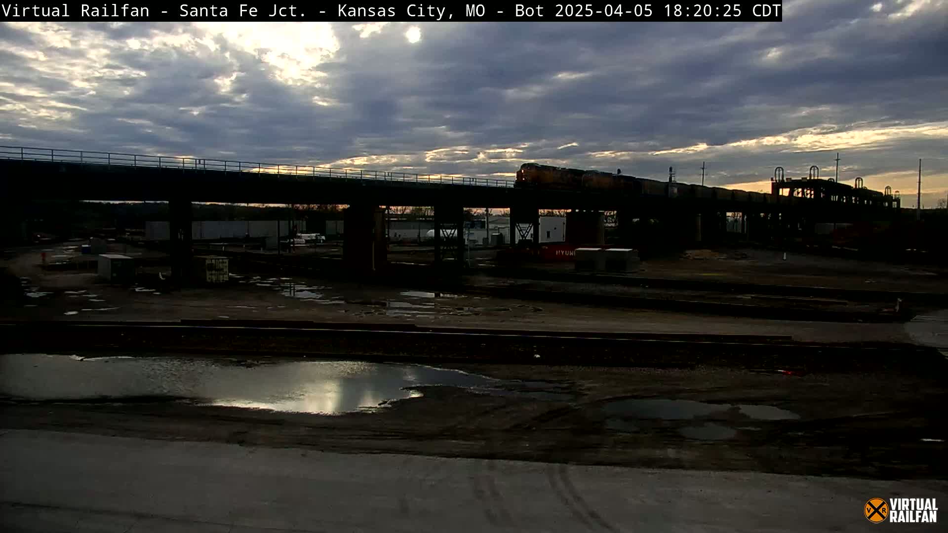 A long freight train crosses a bridge under a partly cloudy twilight sky over a large, mostly empty rail yard with puddles of water on the ground.
