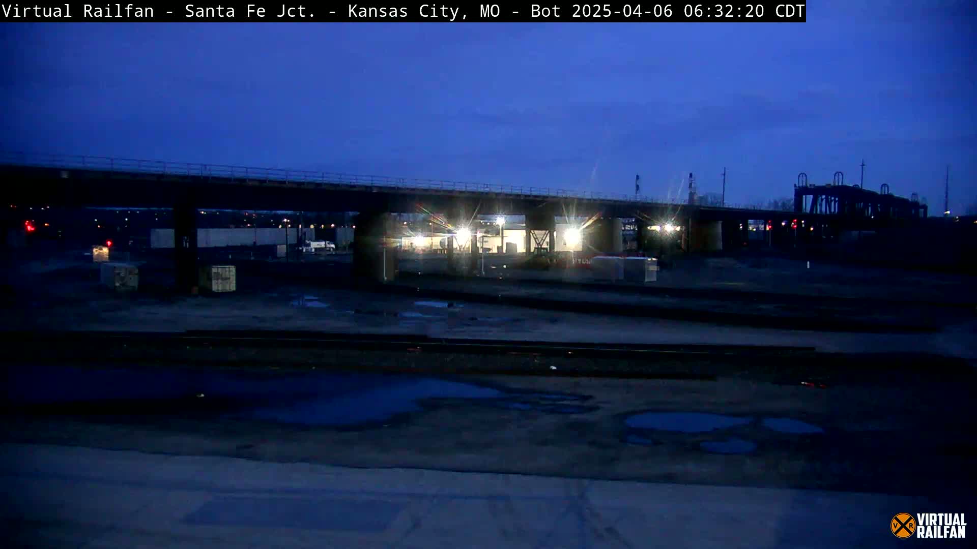 A dark, pre-dawn view of a rail yard with a bridge overhead, showing puddles of water on the ground.