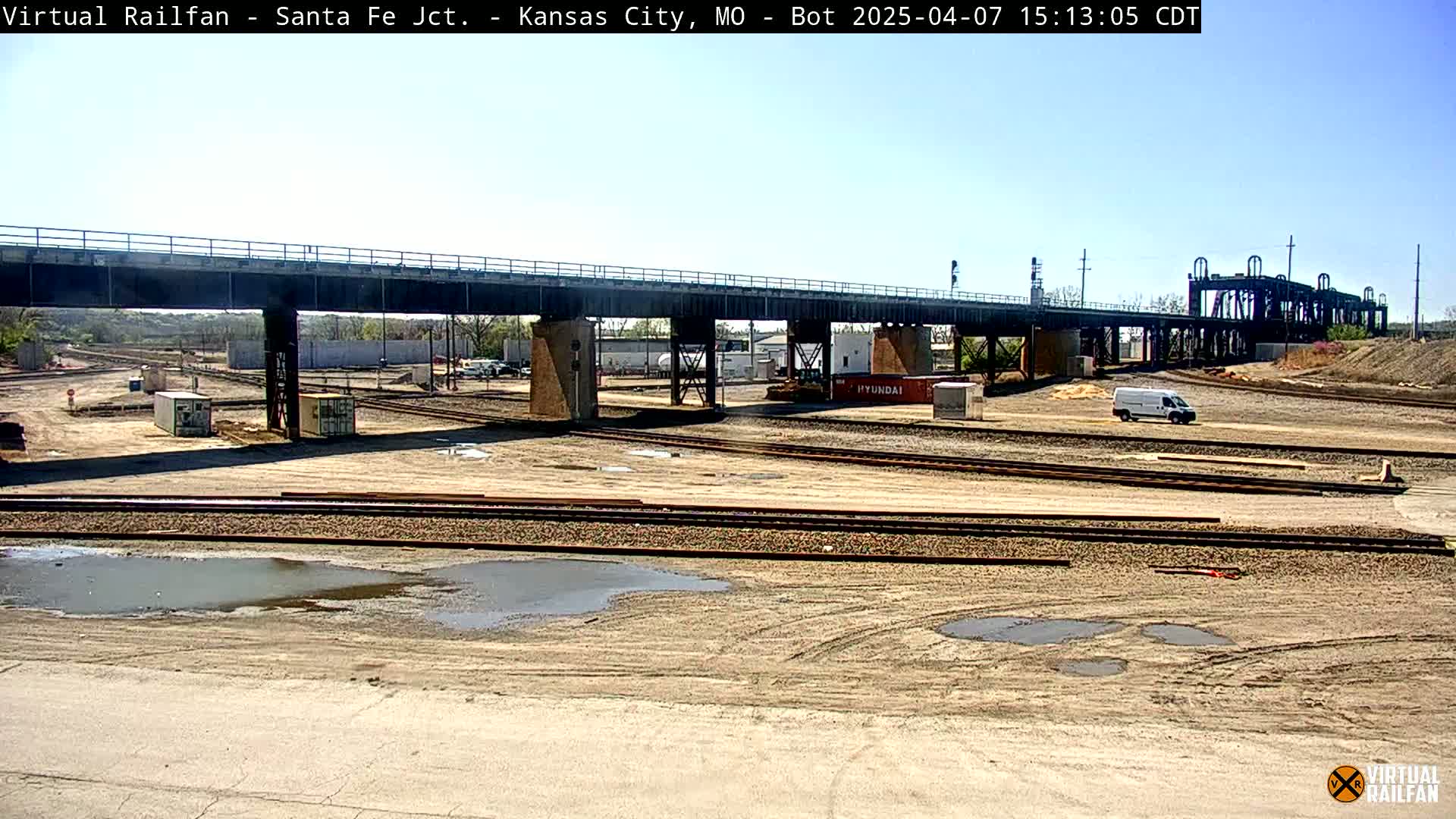A mostly sunny day reveals a train yard under a bridge with a few puddles and some construction equipment.