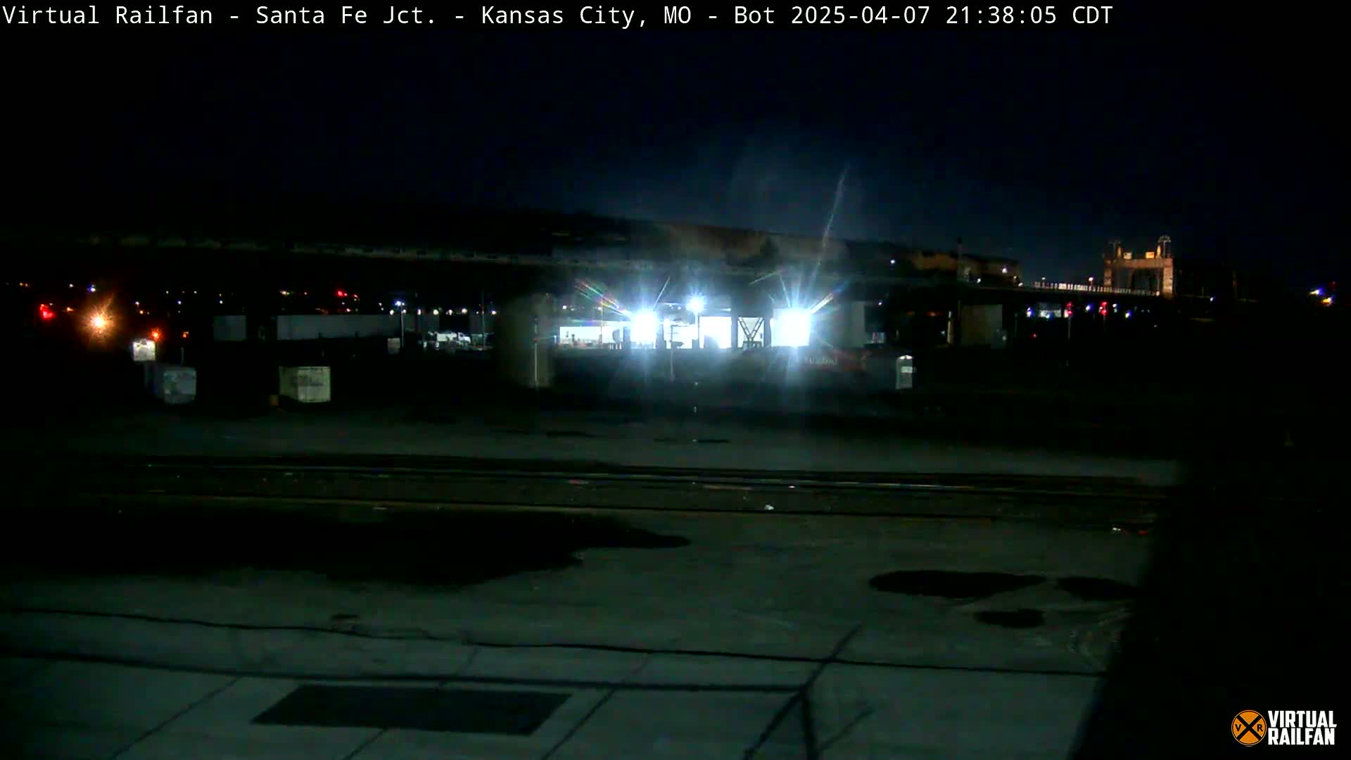 A nighttime view of a train yard with a brightly lit structure in the center, under a dark, clear sky.